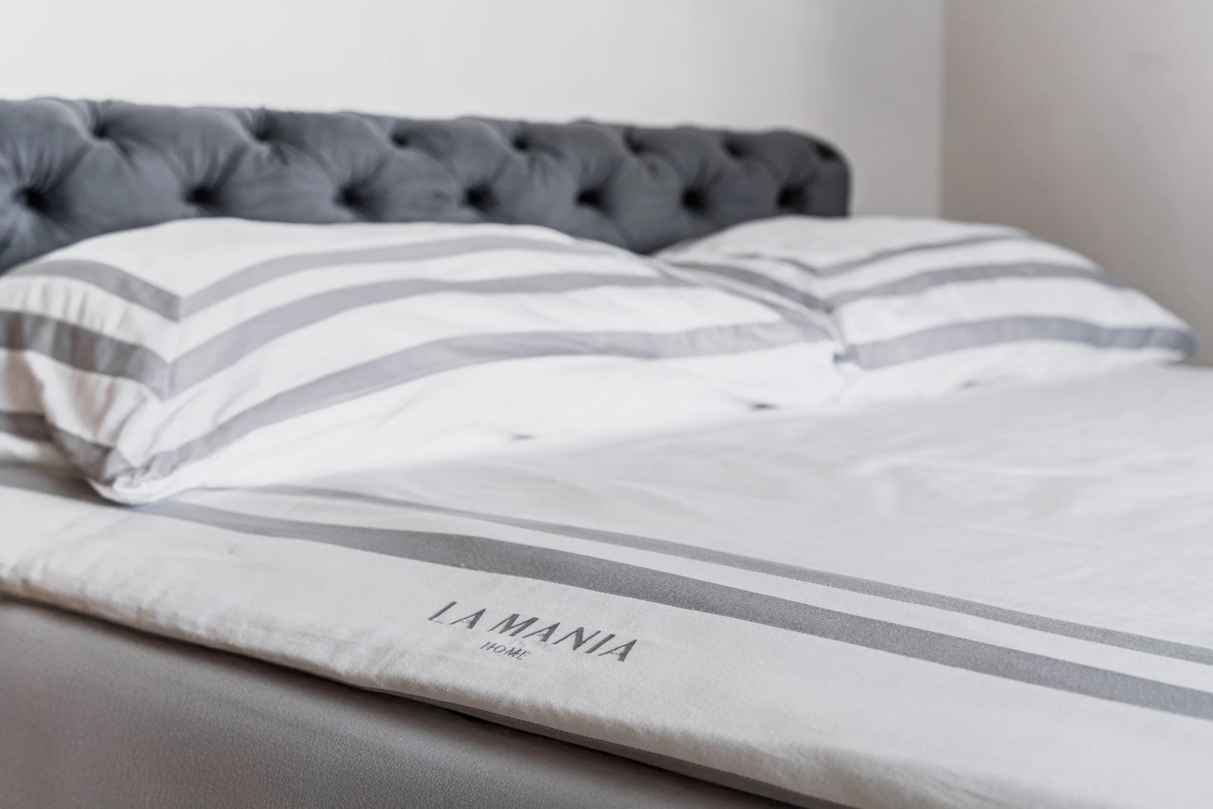 A neatly made bed with a tufted gray headboard, white bed linens, and gray and white striped pillows. The bedspread has the text 'La Manja Home' on it.