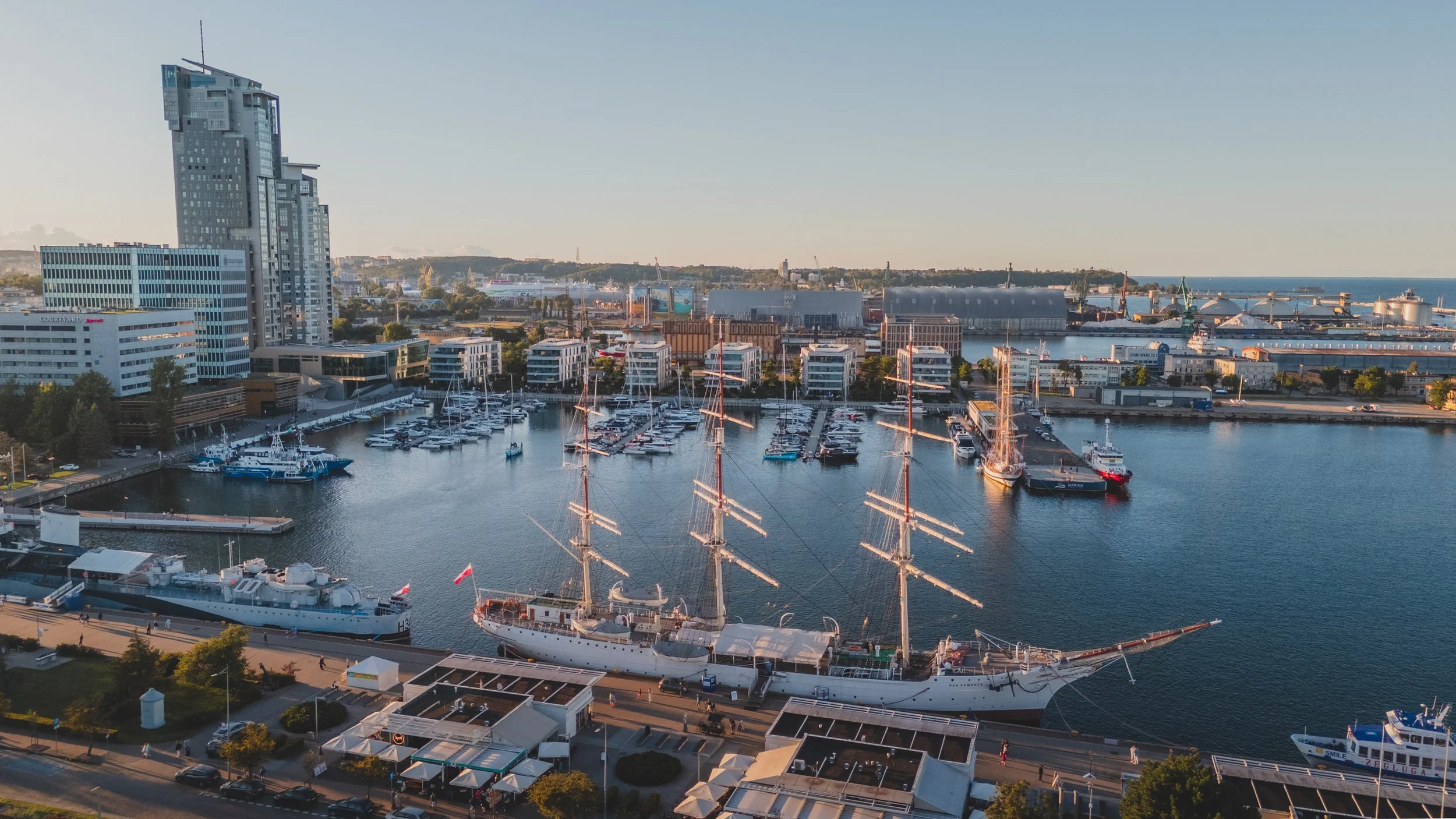 A harbor with sailboats and yachts, including a large sailing ship, in front of a city skyline with modern buildings and a waterfront promenade, under a clear sky.