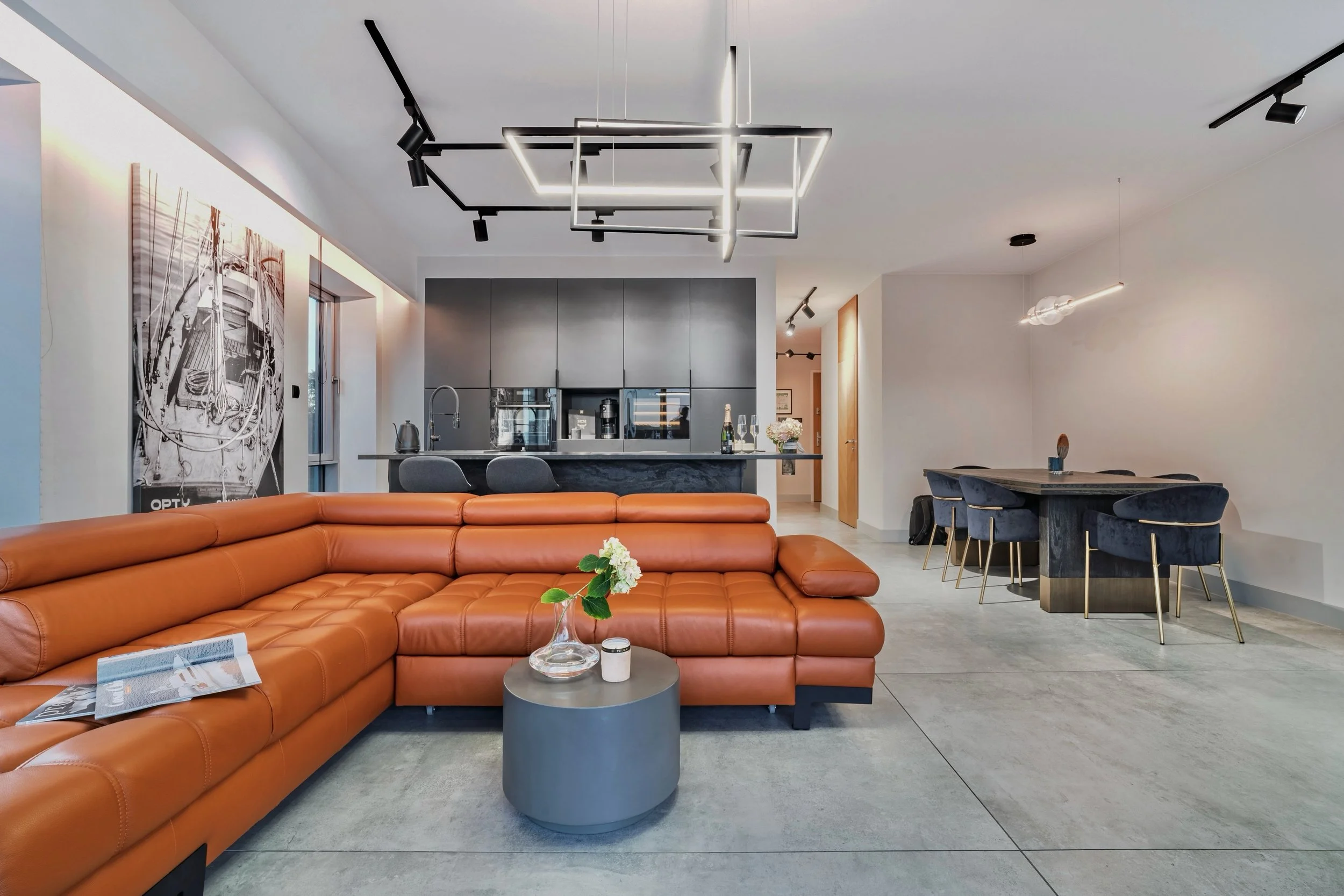 Modern open-concept living space with a brown leather sectional sofa, a gray cylindrical coffee table with a flower vase and candle, an integrated black and gray kitchen with an island, a black dining table with six blue chairs, minimalist lighting f