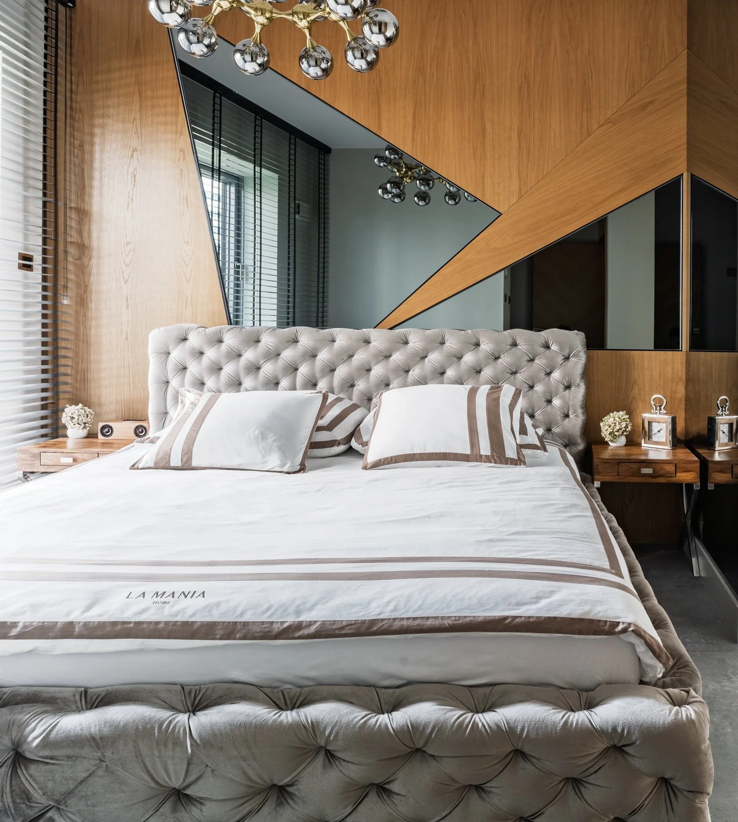 Modern bedroom with a tufted beige headboard, white bed linens with brown accents, and two matching bedside tables with decorative items including small flower arrangements and clock-like objects. A large, geometric mirror and wooden wall paneling are visible in the background.
