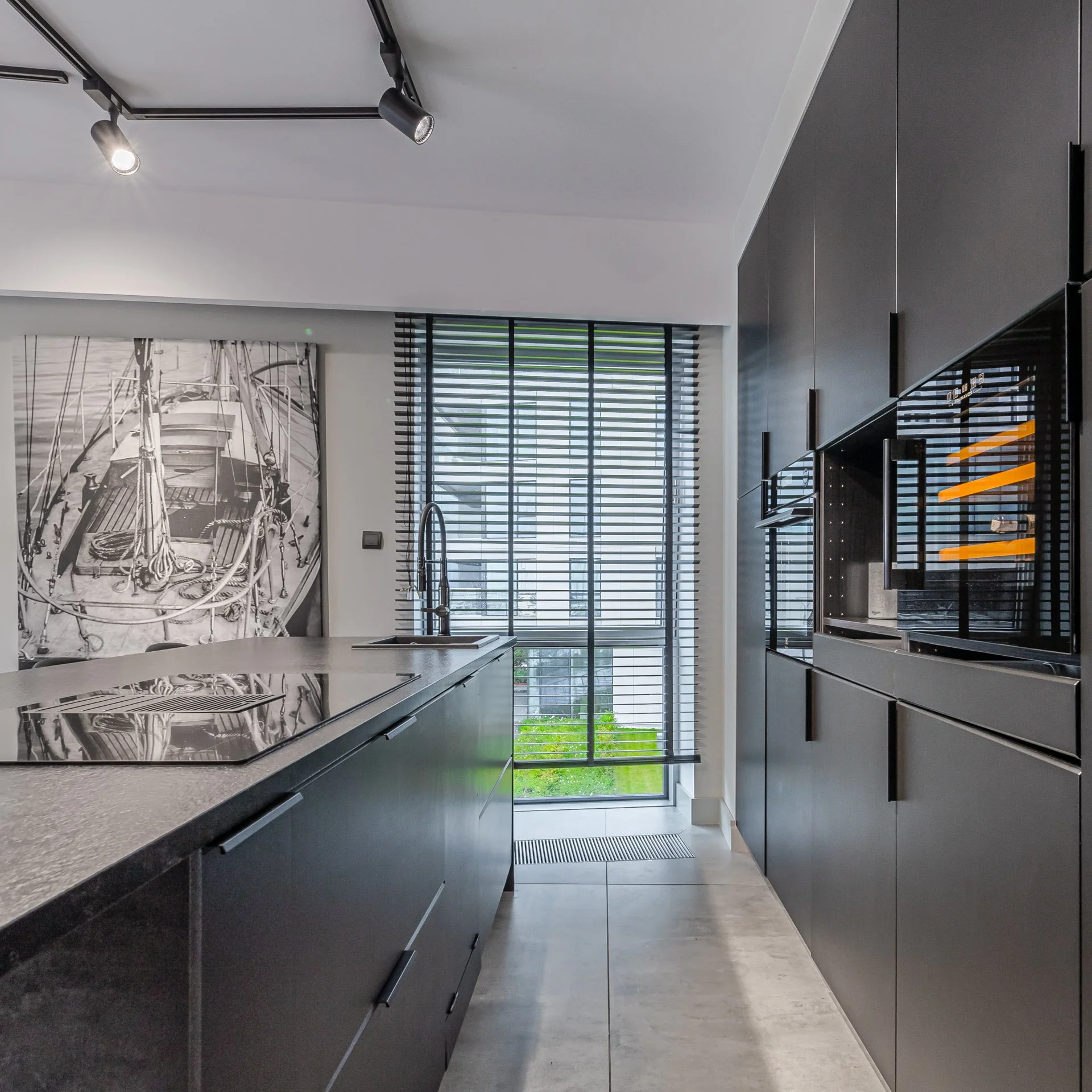 Modern kitchen with black cabinets, a gray countertop, a window with horizontal blinds, and a rear door with glass panels showing a green lawn outside.