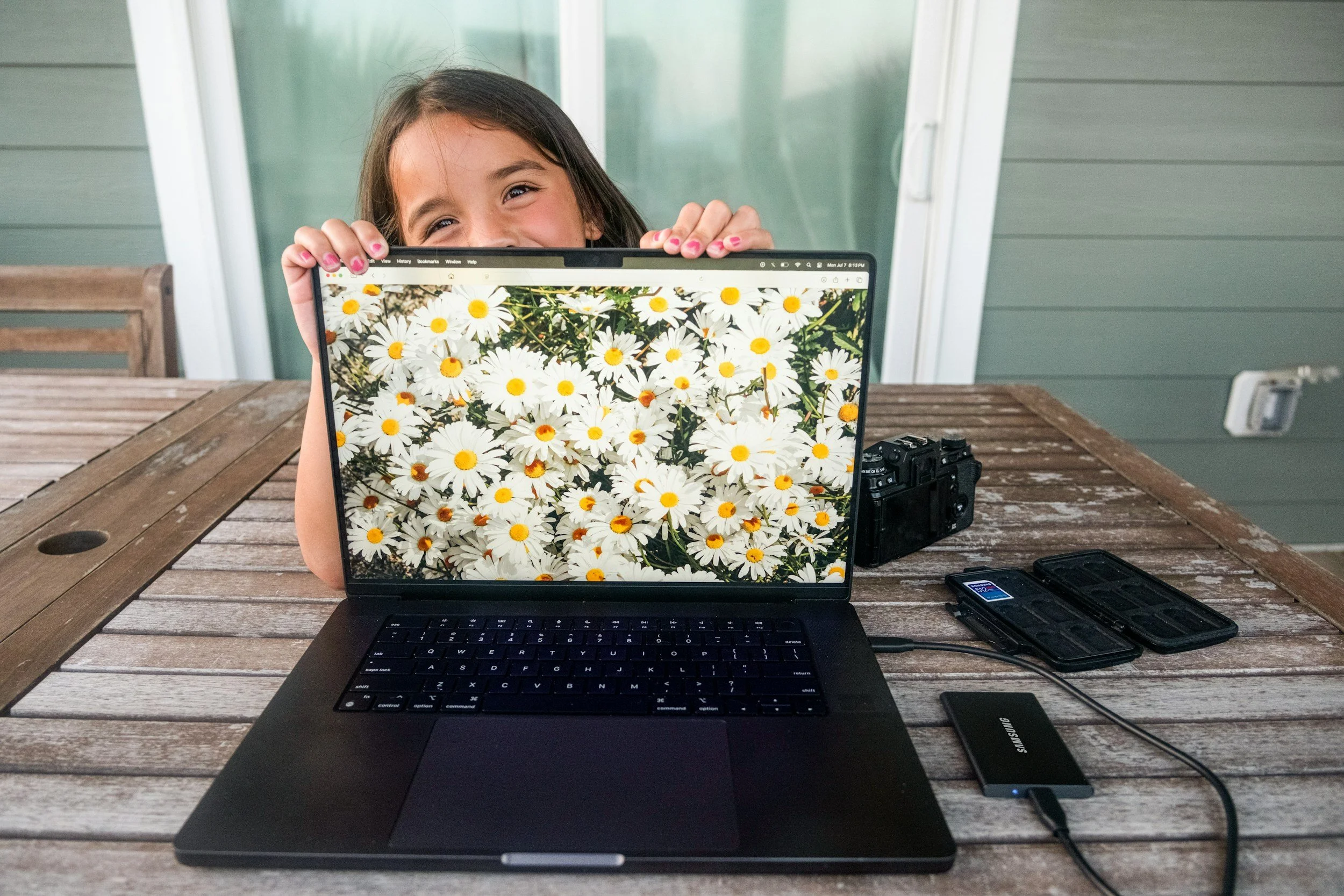 Young girl smiling behind a laptop showing a picture of daisies, on a wooden outdoor table with camera and external drives.