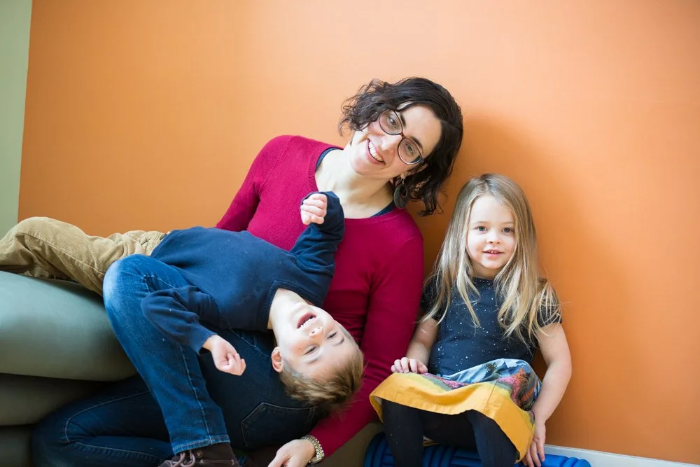A woman with glasses and curly dark hair smiling, sitting on the floor with two children near a colorful wall. One child with light hair and a blue dress is sitting on the woman's lap, smiling. The other child with brown hair in a navy hoodie and khaki pants is lying on the floor with head on the woman's lap, looking up.