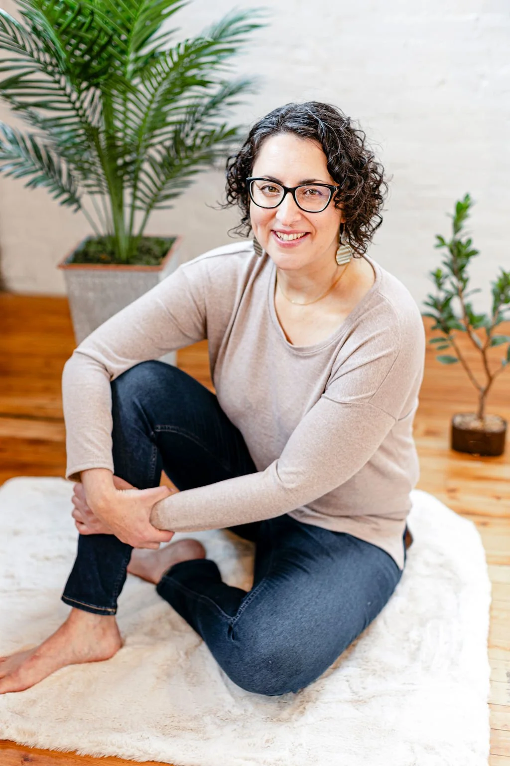 A woman with curly dark hair and glasses sitting on a white rug, smiling at the camera, with two potted plants in the background in a room with wooden flooring.