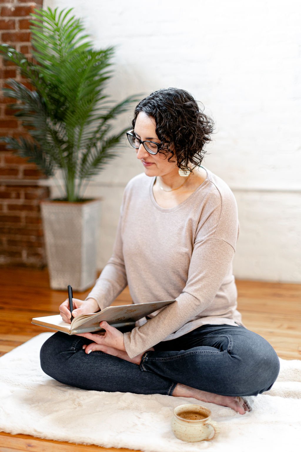 A woman with curly hair and glasses sitting cross-legged on a soft rug, writing in a notebook, with a potted plant and coffee mug nearby.