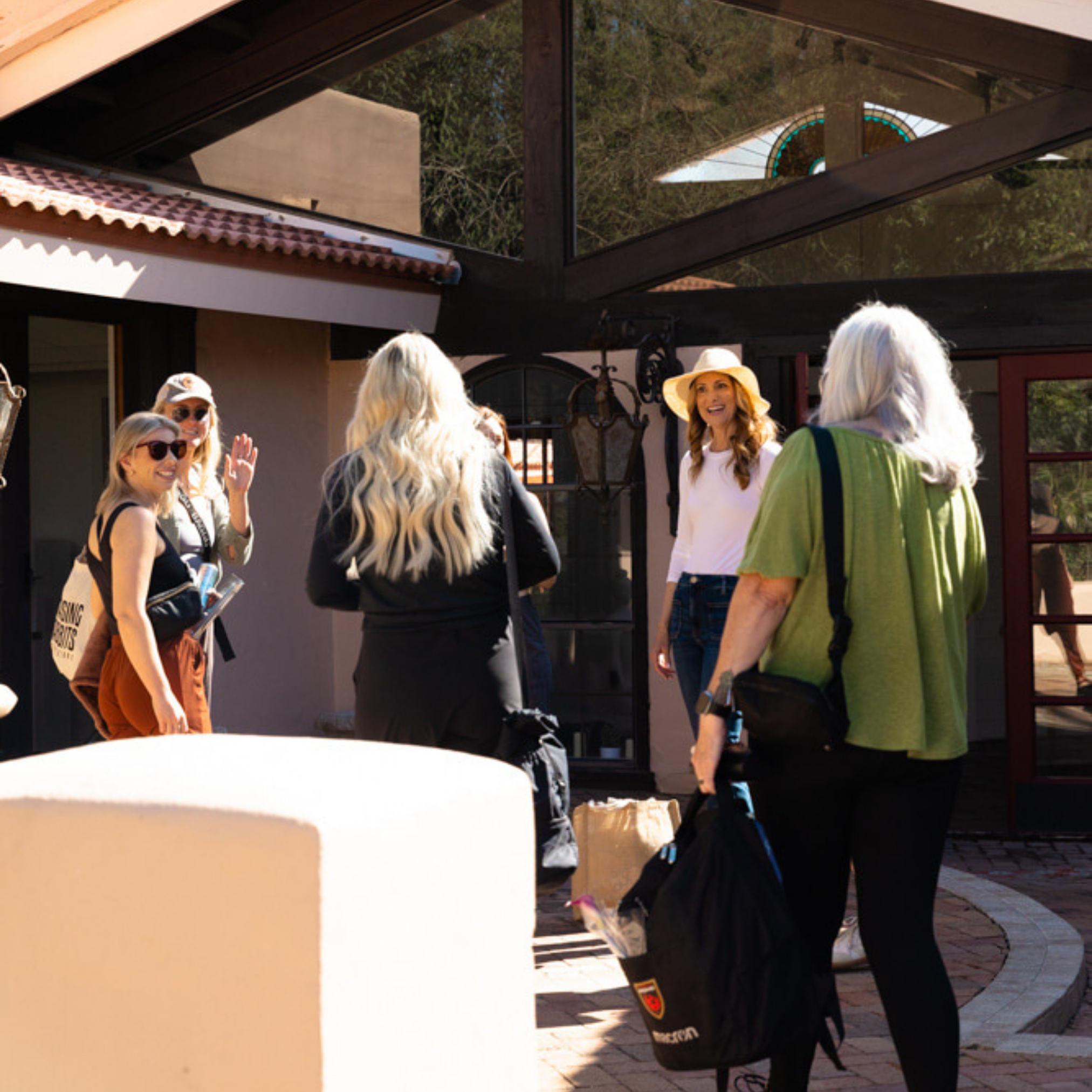 Five women gather outside a building, some carrying bags, while one woman smiles and waves near the entrance for retreat planning support.