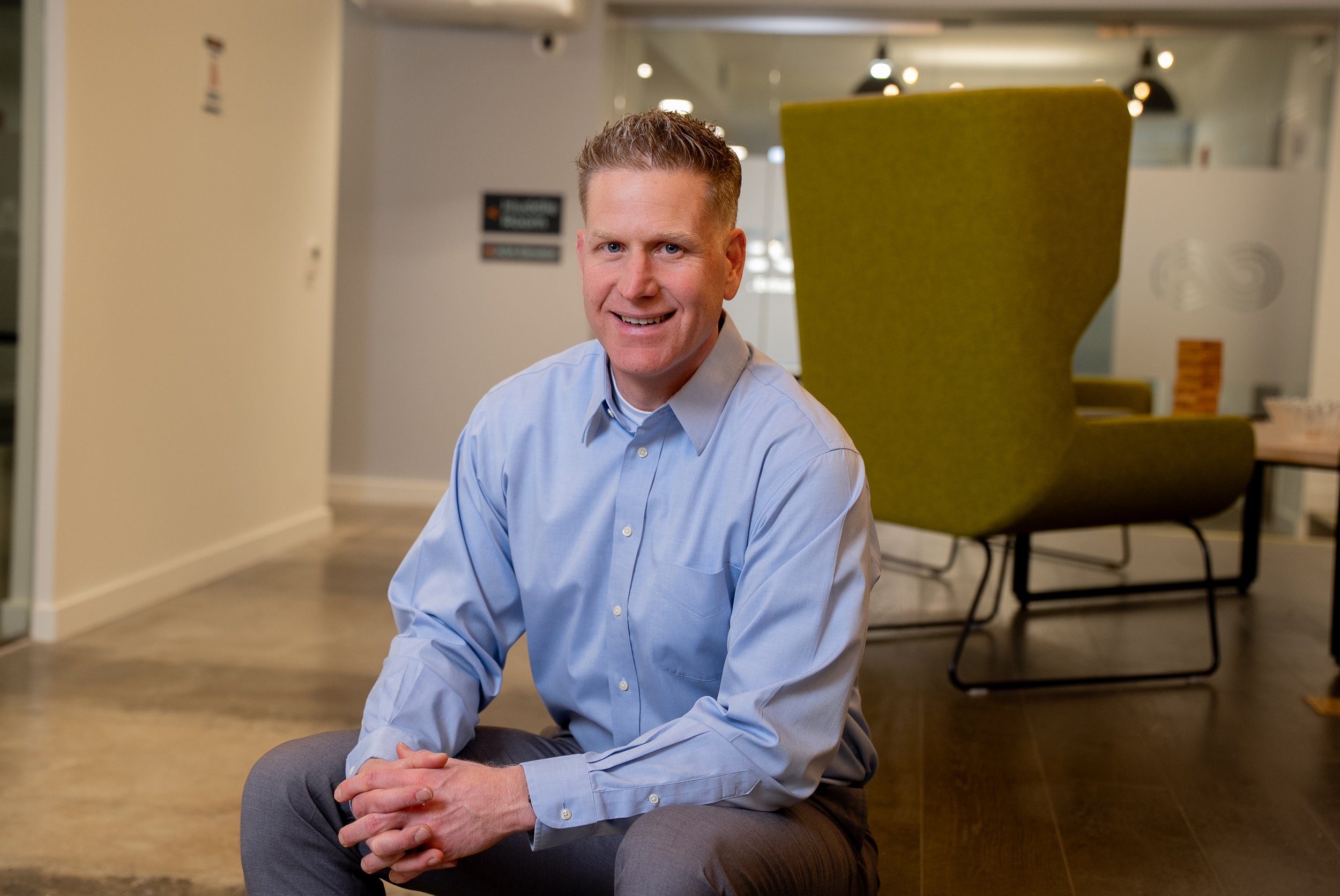 A photo of Michael, owner of Crosswind CFO, A smiling man in a light blue dress shirt sitting on the floor of an office lounge area.