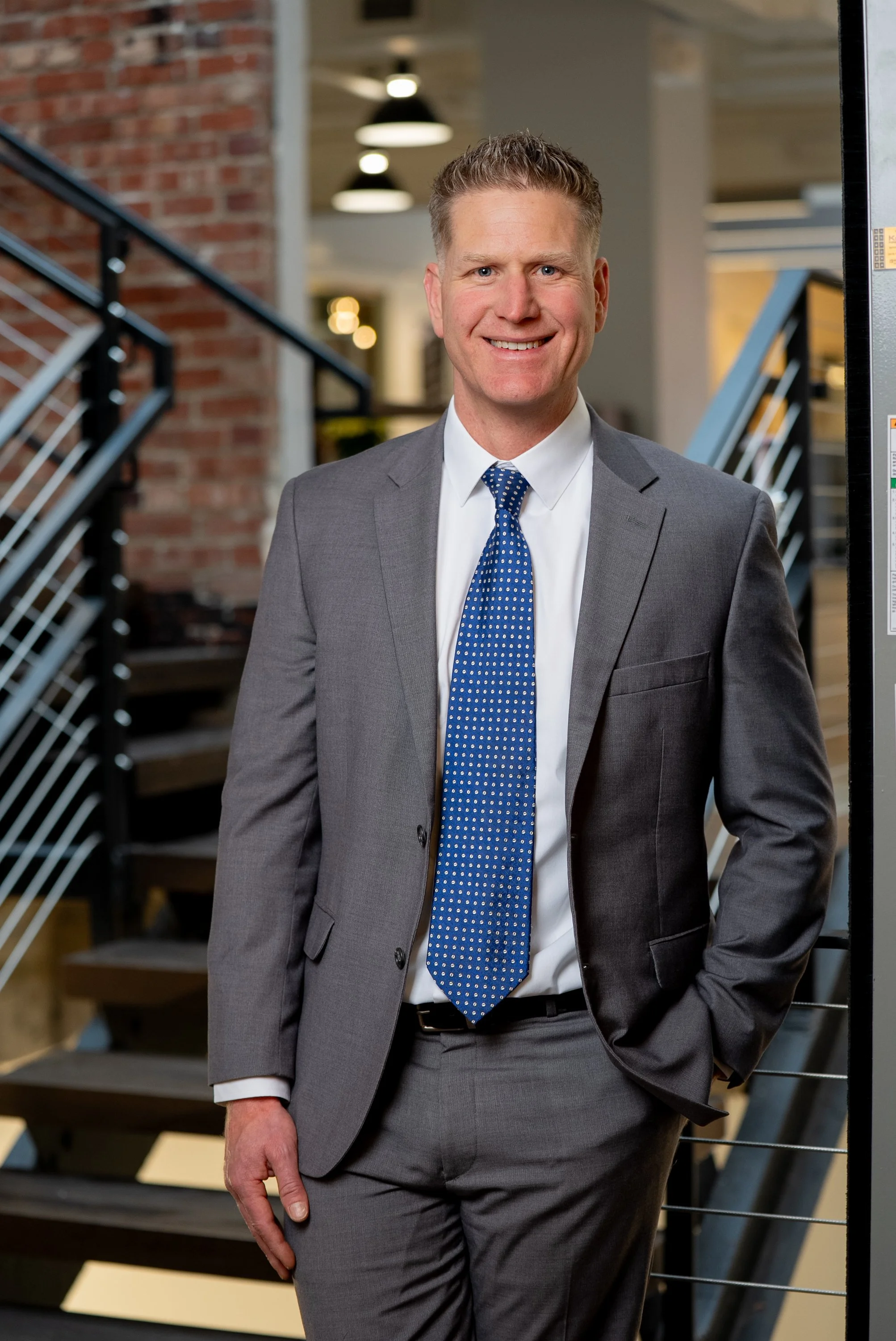 A photo of Michael, owner of Crosswind CFO, A man in a gray suit and blue tie standing indoors near a staircase, smiling at the camera.