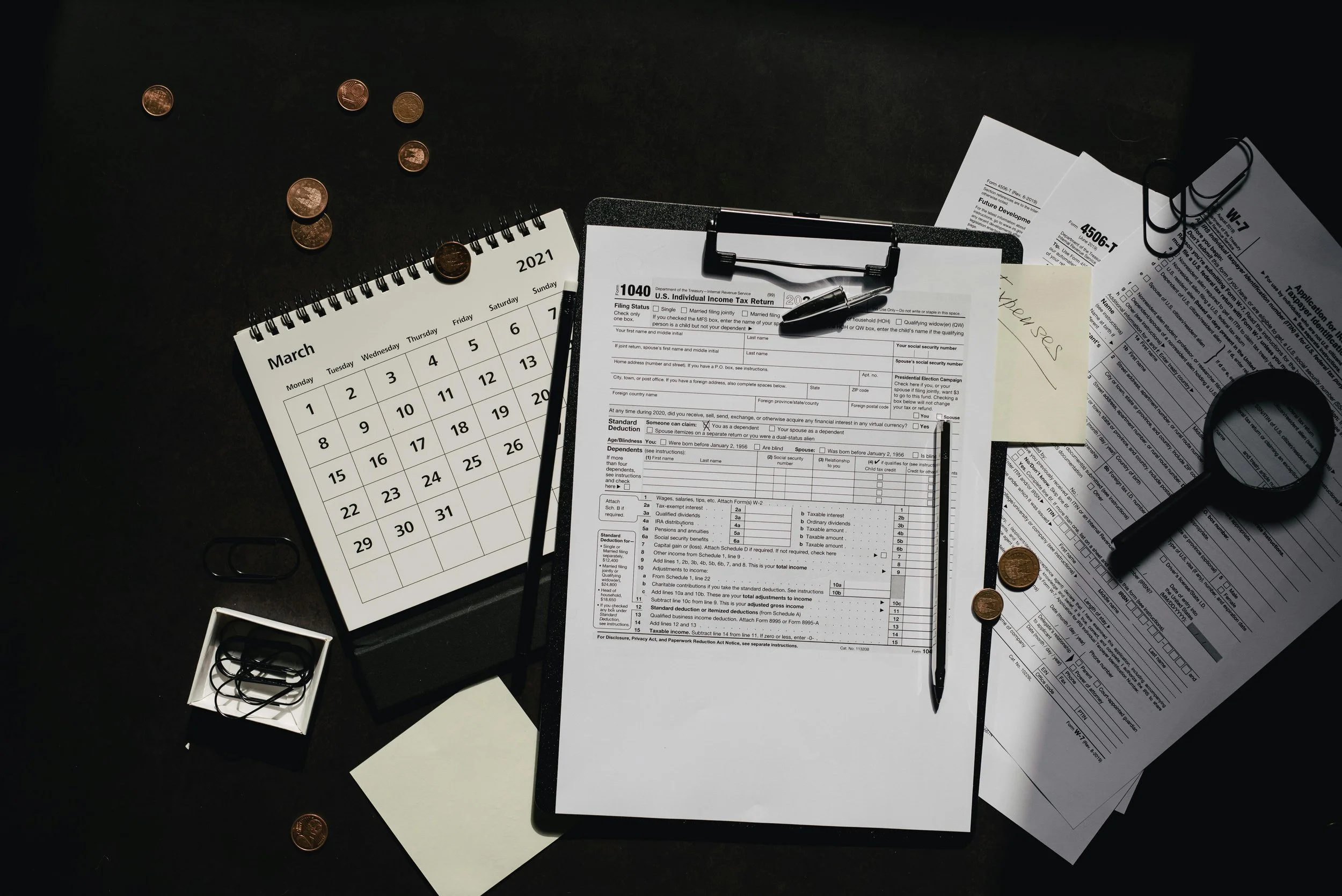Desk cluttered with a 2021 calendar, tax forms, a pen, eyeglasses, a small box with earbuds, and scattered pennies.
