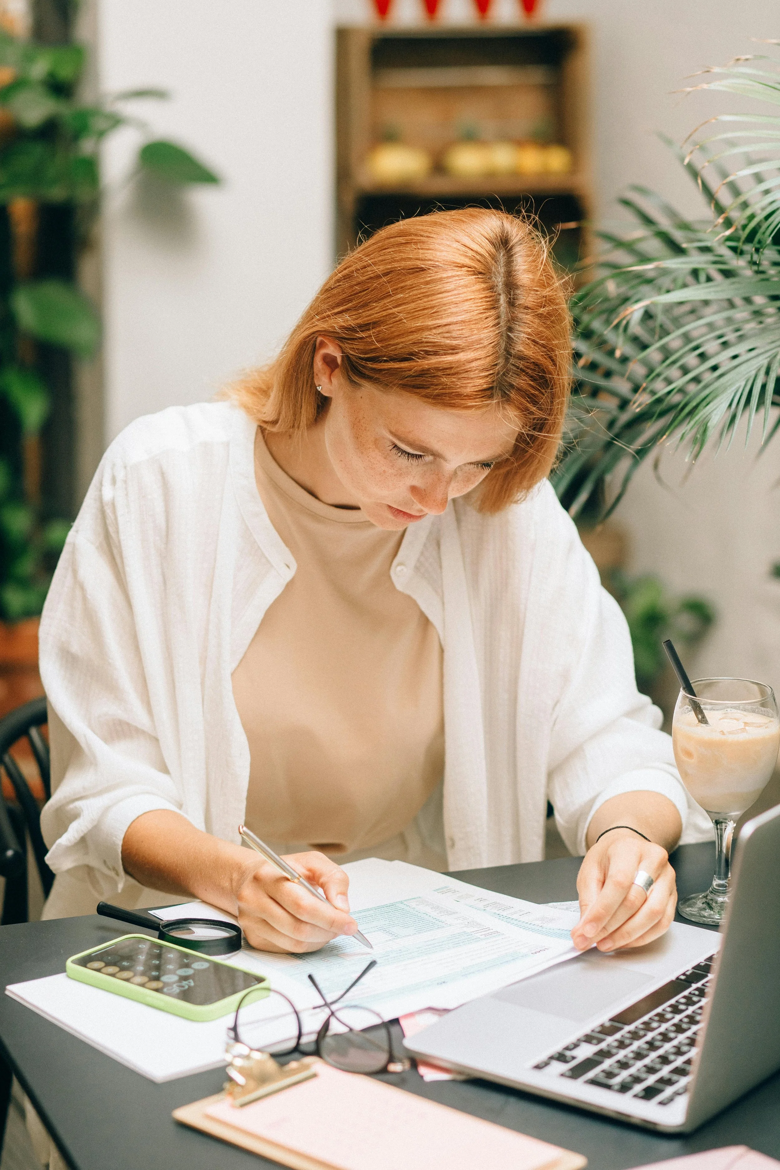 A woman with short, red hair and freckles, wearing a beige top and a white shirt, sitting at a desk writing on a document.

On her desk are sunglasses, a calculator, a smartphone with a green case, and a laptop. There is a calculator and a pair of glasses on the desk.

In the background, there are plants and a wooden shelf with some objects.