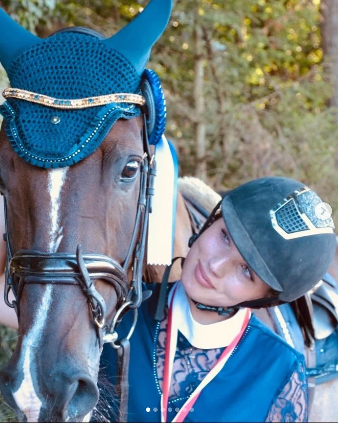 Junge Frau mit Helm und blauer Reiterjacke neben einem braunen Pferd mit blauer Turniermütze im Freien.