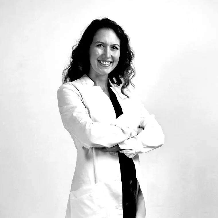 A woman smiling with arms crossed, wearing a white lab coat against a plain background.