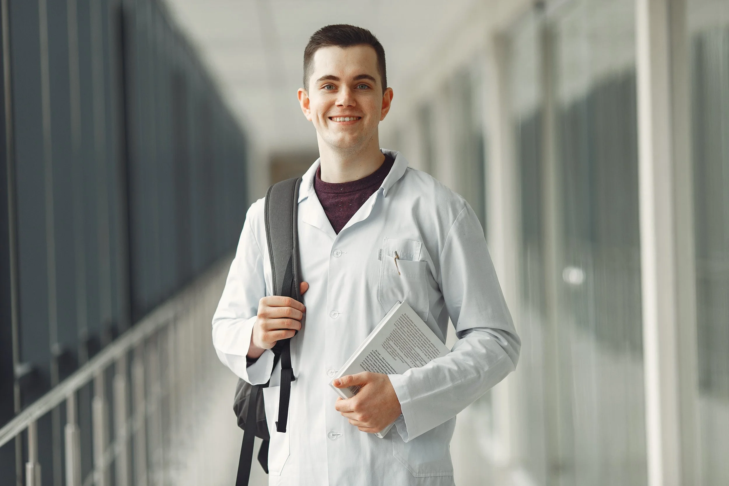 A young man in a white medical coat holding books and a backpack, standing and smiling in a modern corridor with glass walls.