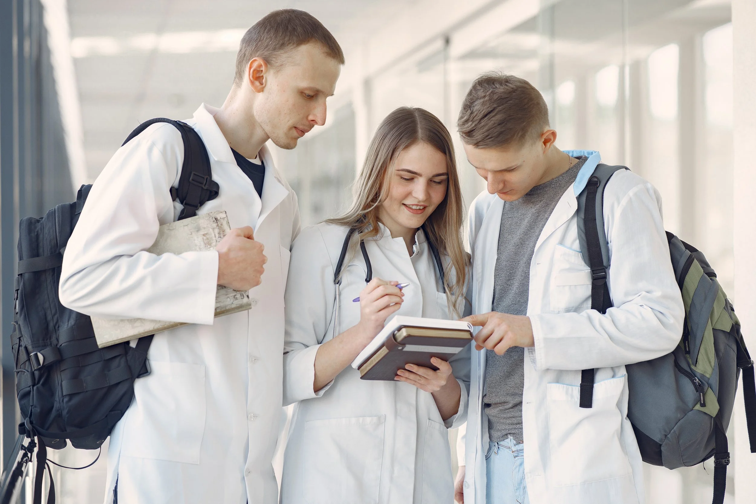 Three medical students with backpacks, wearing white lab coats, standing together in a bright hallway, looking at a tablet device held by one of them and discussing.