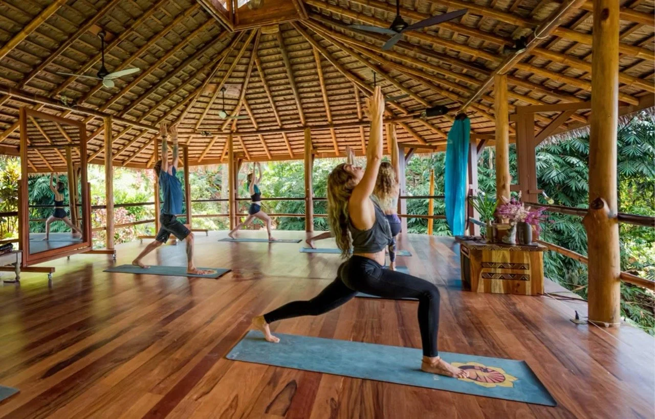 Group of people practicing yoga on mats inside a wooden, open-air pavilion surrounded by lush greenery.