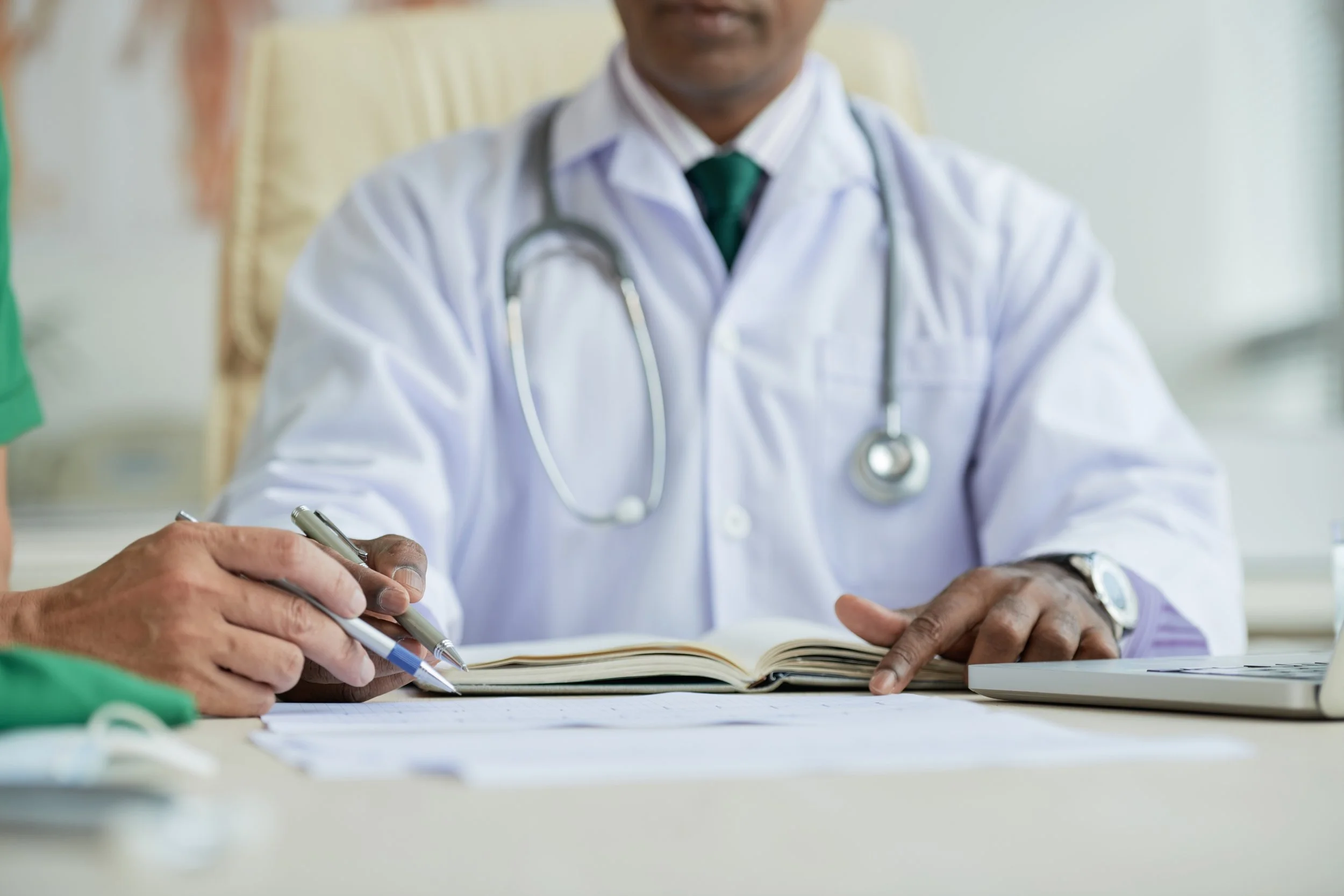 A doctor wearing a white coat and stethoscope sitting at a desk, writing in a notebook with another person holding a pen nearby.