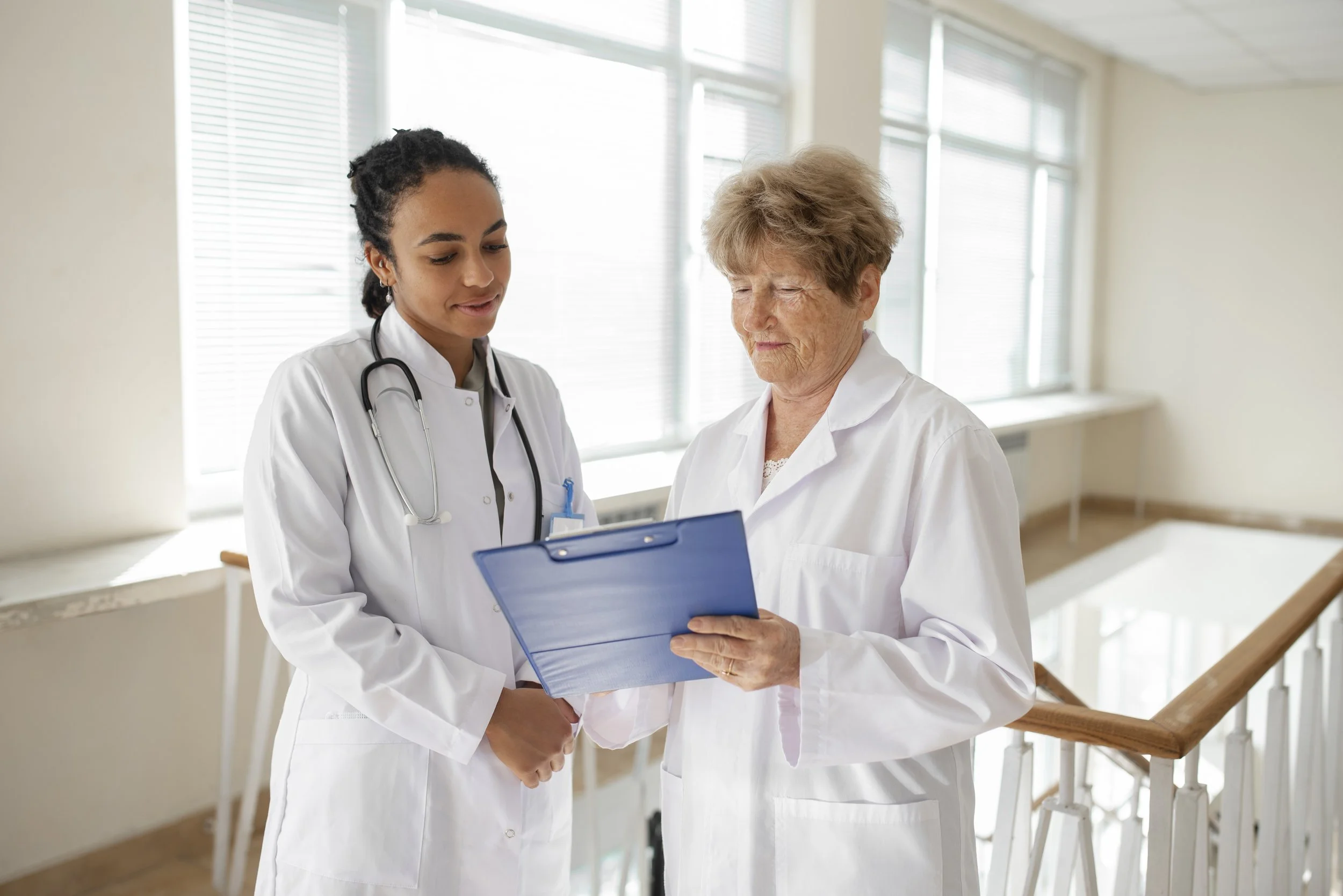 Two female medical professionals, one young and one older, standing together in a hospital corridor, reviewing a blue clipboard.