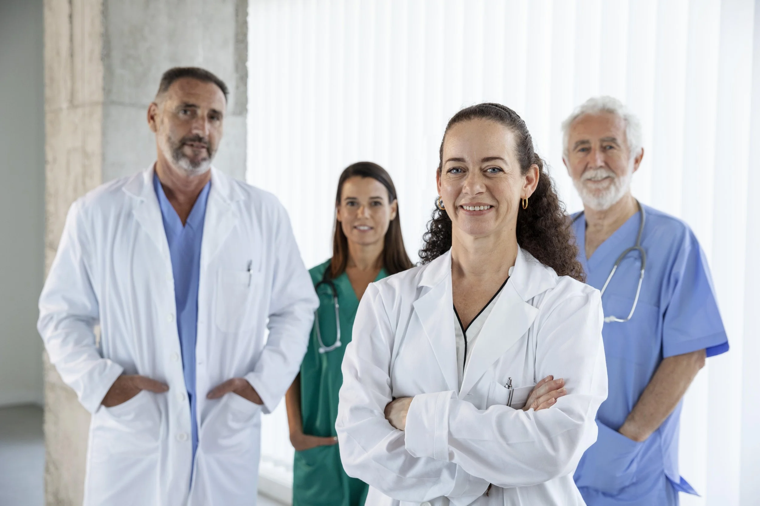 Group of four healthcare professionals in a clinic or hospital, with a woman doctor in the foreground smiling and with arms crossed, and three other medical staff standing behind her.