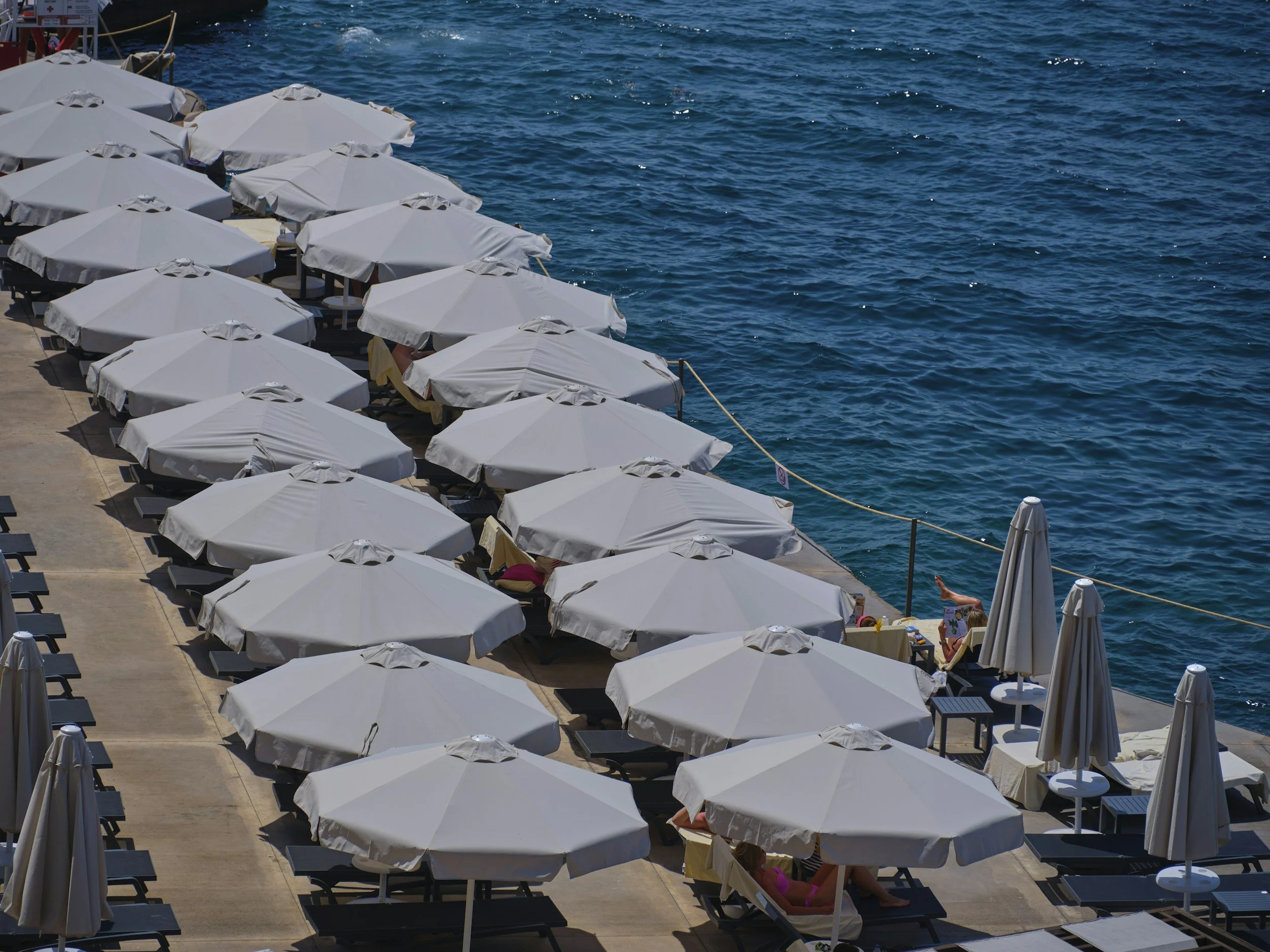 Beach chairs and umbrellas lined up along a pier by the water with people relaxing and swimming in the background.