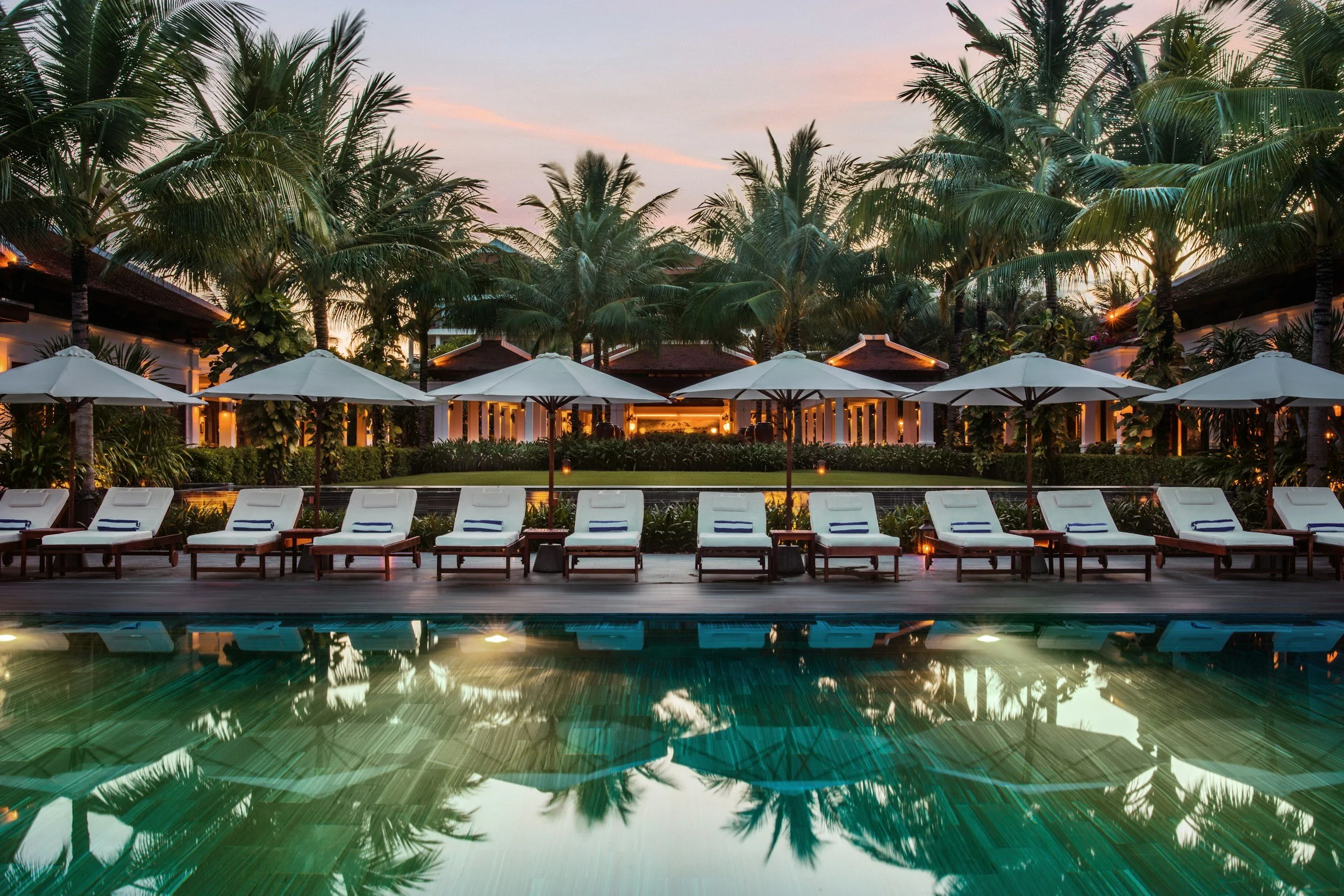 Luxury resort pool area at sunset with white lounge chairs and umbrellas, surrounded by palm trees and lush greenery.