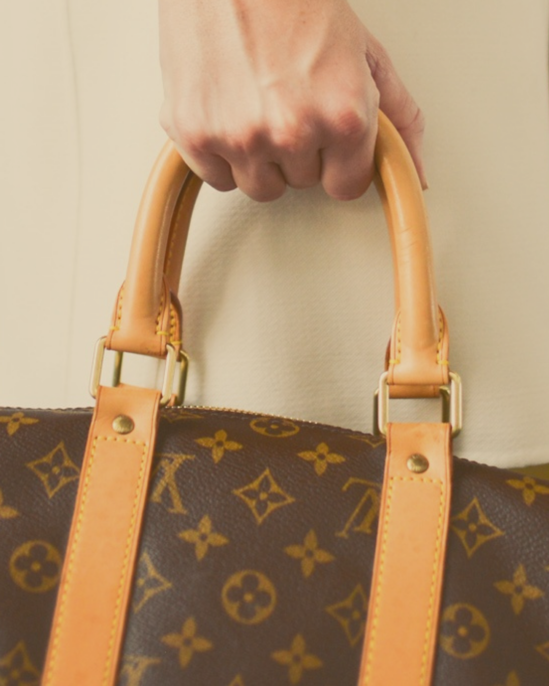 Close-up of a hand holding the handle of a Louis Vuitton handbag with the classic monogram pattern and tan leather straps.