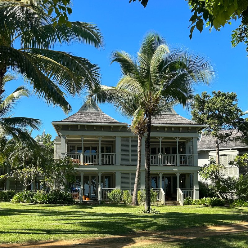 Two-story house with a grey exterior, balconies on both floors, surrounded by palm trees and greenery, under a clear blue sky.