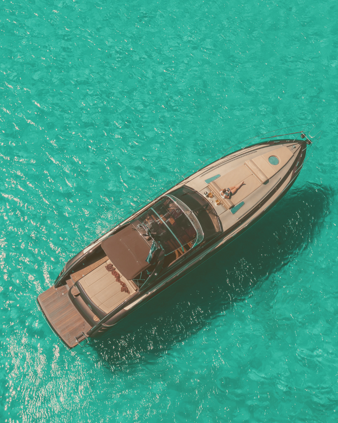 Aerial view of a luxury yacht with a person relaxing on the sun deck, floating on clear turquoise water.