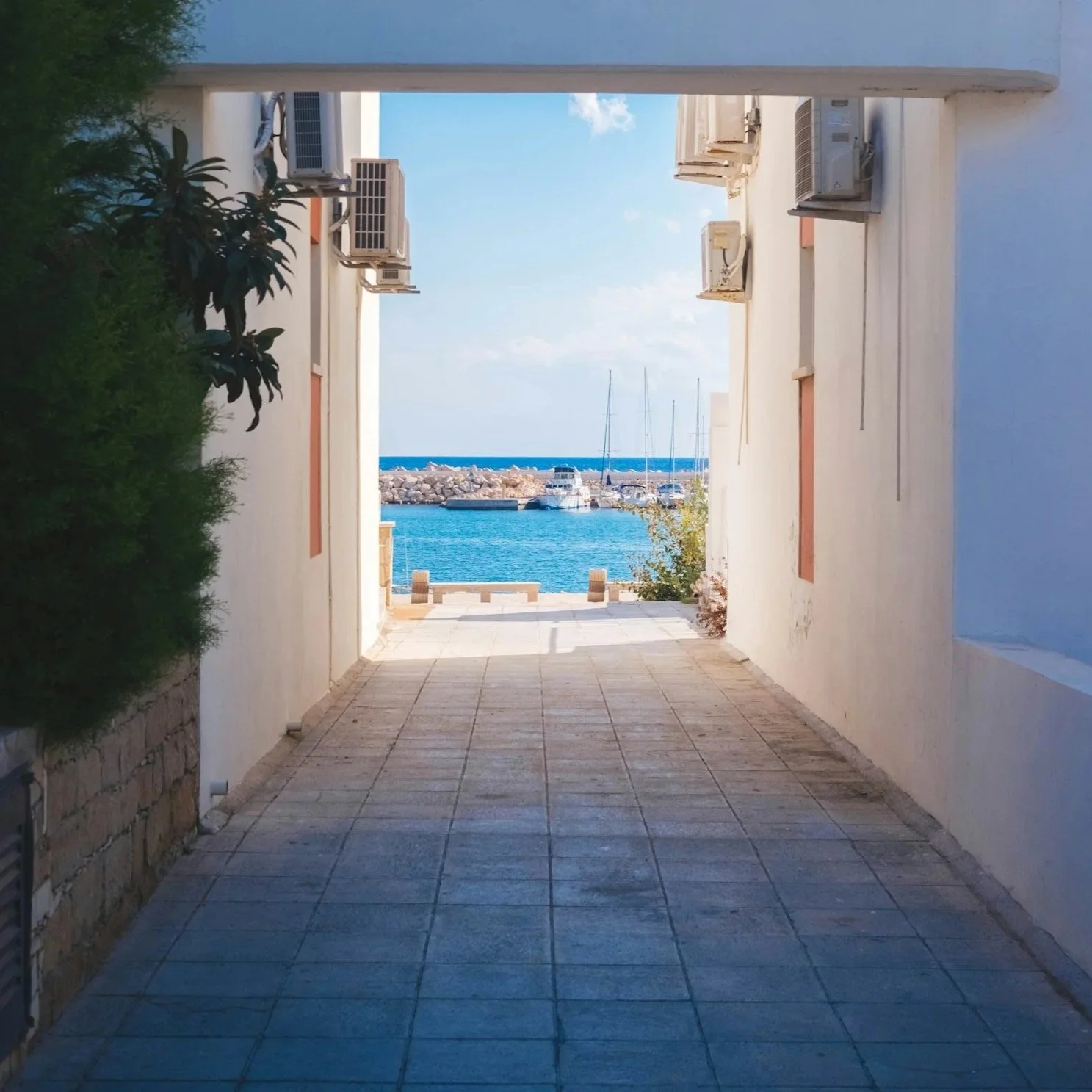 A narrow alleyway with white walls on both sides, leading to a view of a marina with boats and yachts on the water, and a breakwater with rocks in the distance under a blue sky.
