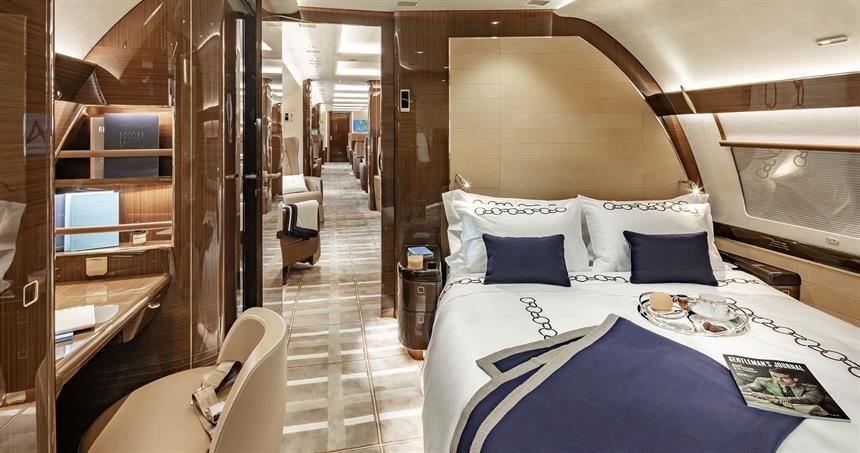 Luxurious airplane bedroom with a large bed, navy and white bedding, and seating area, wood paneling, and a corridor with seats and amenities in the background.
