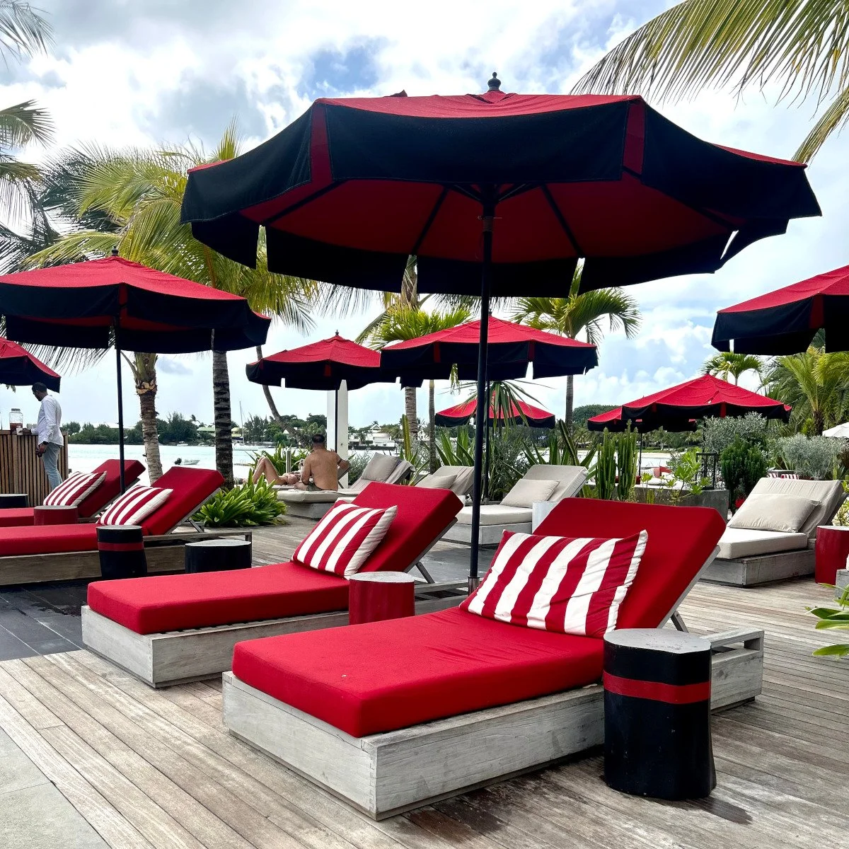 Poolside lounge chairs with red cushions and striped pillows under red and black umbrellas, with palm trees and a body of water in the background.