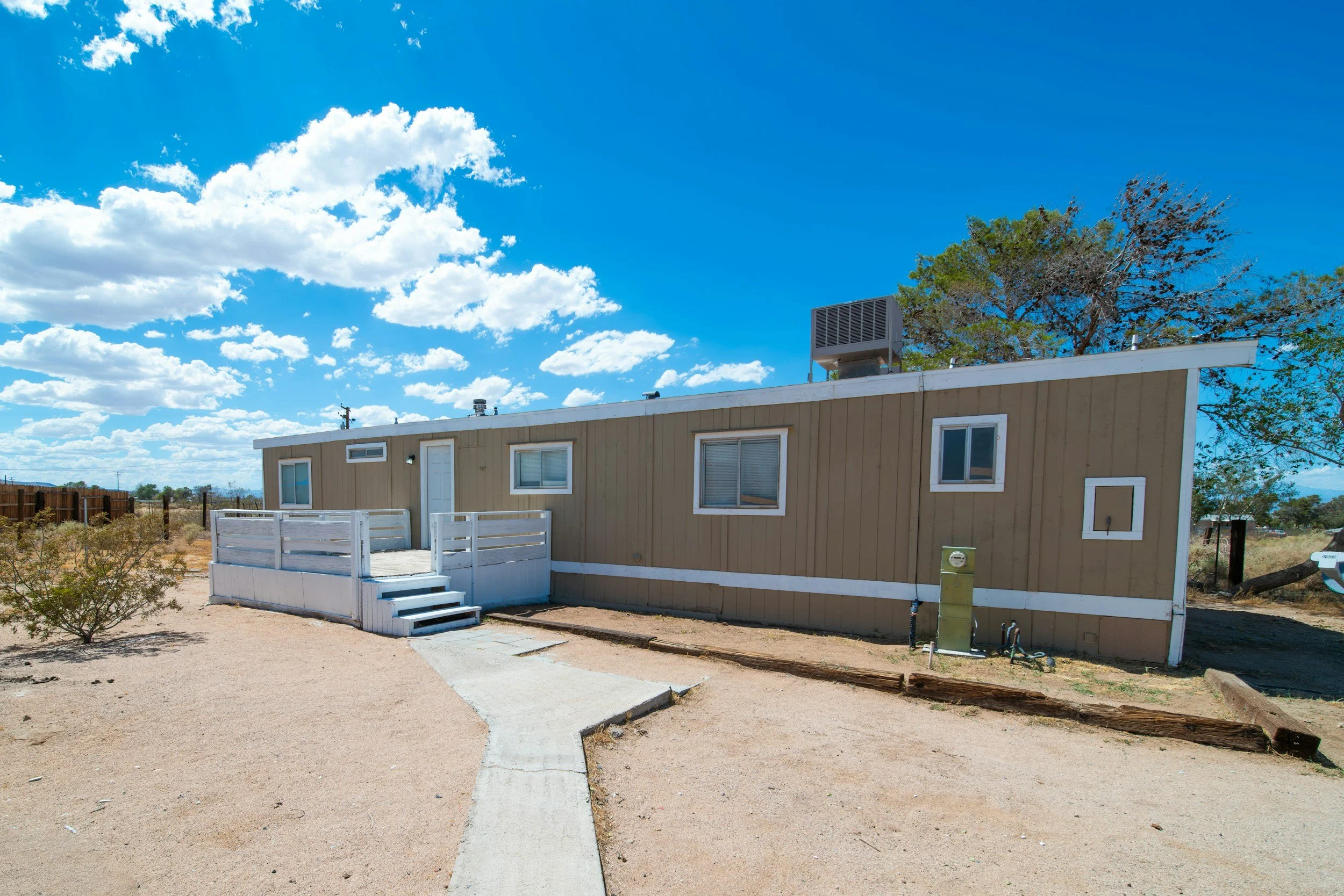 A beige mobile home with a white porch and steps, set in a desert landscape under a bright blue sky with clouds, with trees and a fence in the background.