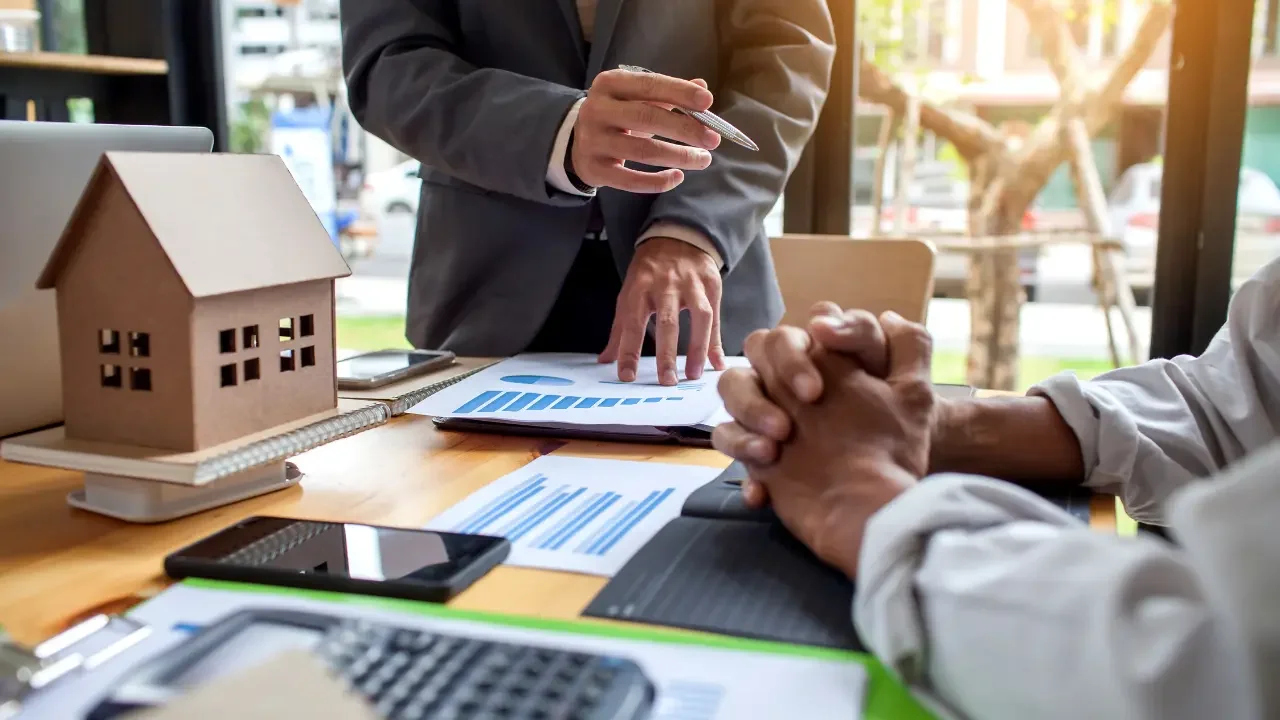 Two people in a meeting, one in a suit and the other in a white shirt, discussing documents on a table with a small house model and a smartphone.