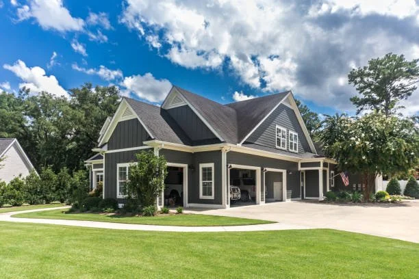 A modern gray house with a three-car garage, surrounded by a well-maintained lawn and trees, under a partly cloudy sky.