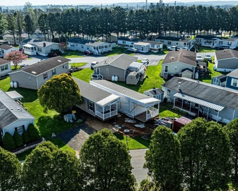 An aerial view of a residential neighborhood with multiple houses, some under construction, surrounded by green lawns and trees, with a line of trees in the background.