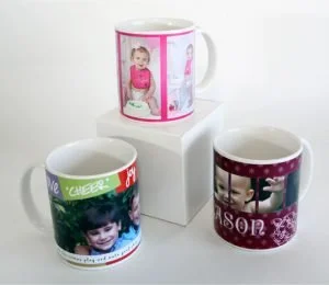 Three ceramic mugs decorated with children's photos and festive designs, displayed on a white pedestal against a plain background.