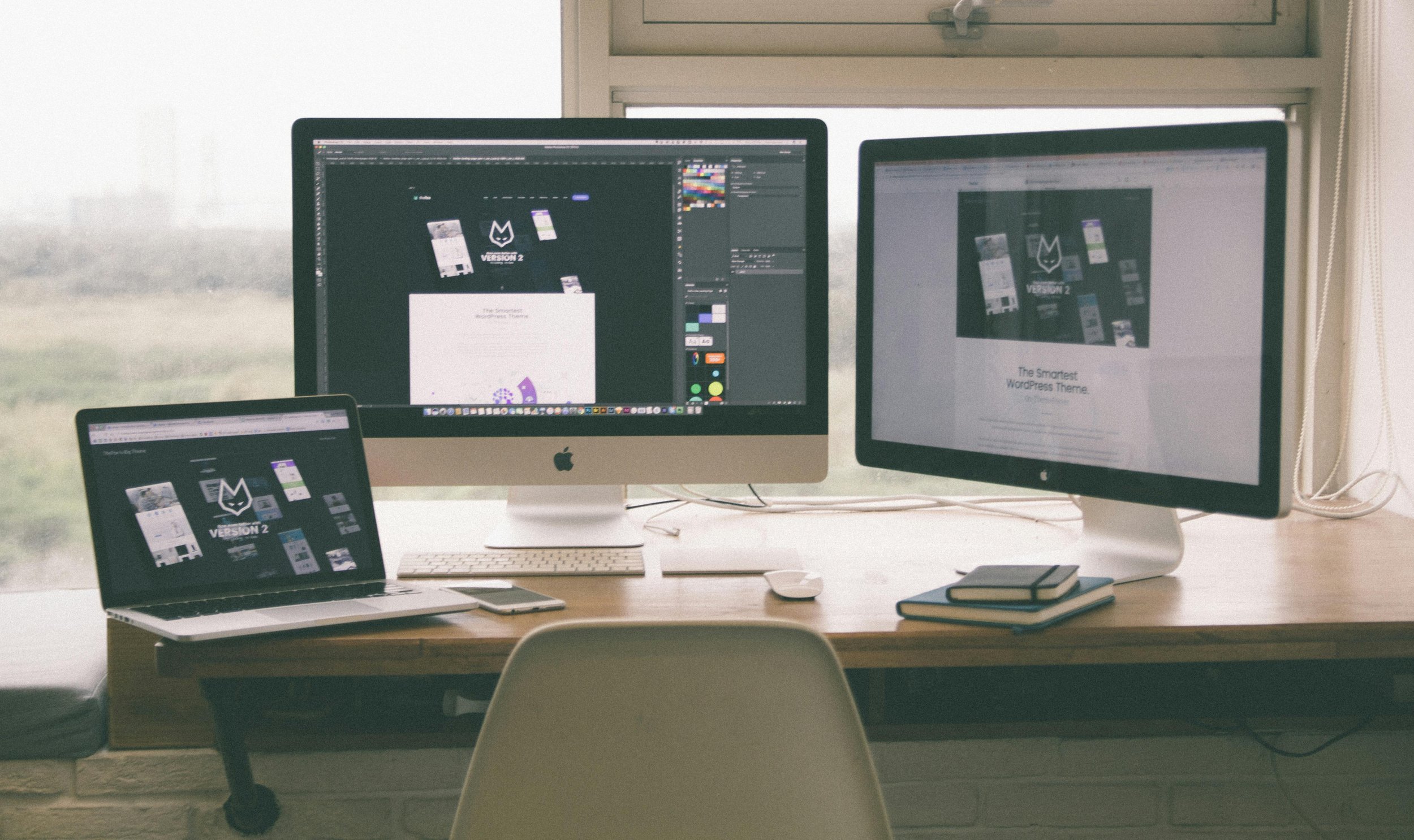A workspace with three screens displaying design projects, an Apple iMac desktop computer, a MacBook laptop, and a large monitor on a wooden desk, near a window with natural light.