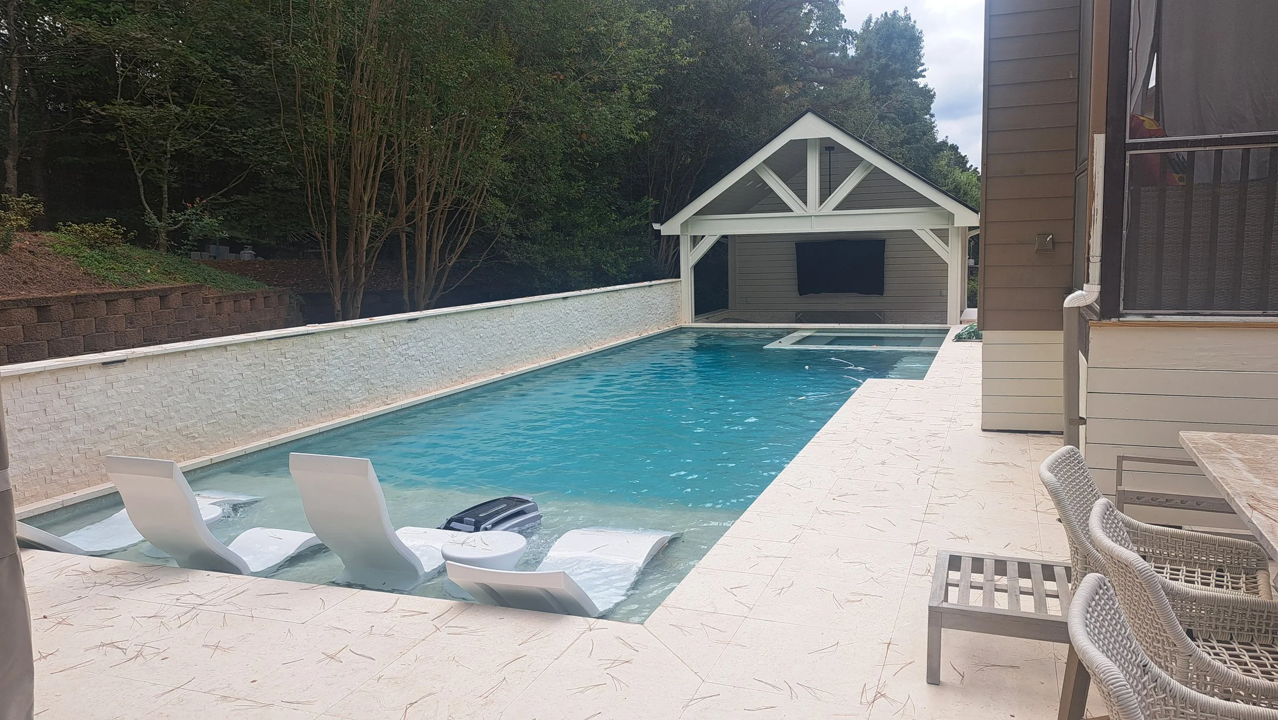 A backyard swimming pool with three white lounge chairs partially submerged in the shallow end. There is a poolside table, and a dining area with wicker chairs. A small enclosed outdoor TV area is visible at the far end of the pool, with a house on the right side of the image and a wooded area in the background.