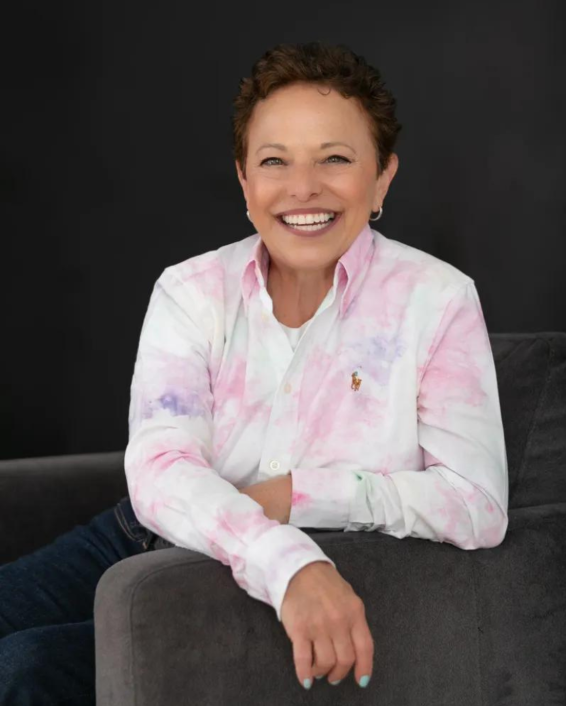 Smiling woman with short brown hair sitting on gray couch against a dark background, wearing a pastel pink and white tie-dye button-up shirt.
