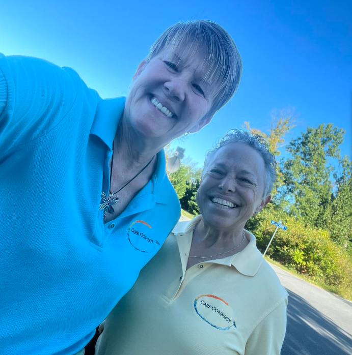 Two smiling women taking a selfie outdoors on a sunny day, wearing shirts with 'Care Connect' logos, with trees and a blue sky in the background.
