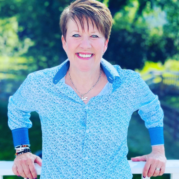 A woman with short brown hair smiling outdoors, wearing a blue patterned shirt with blue cuffs, standing in front of green foliage.