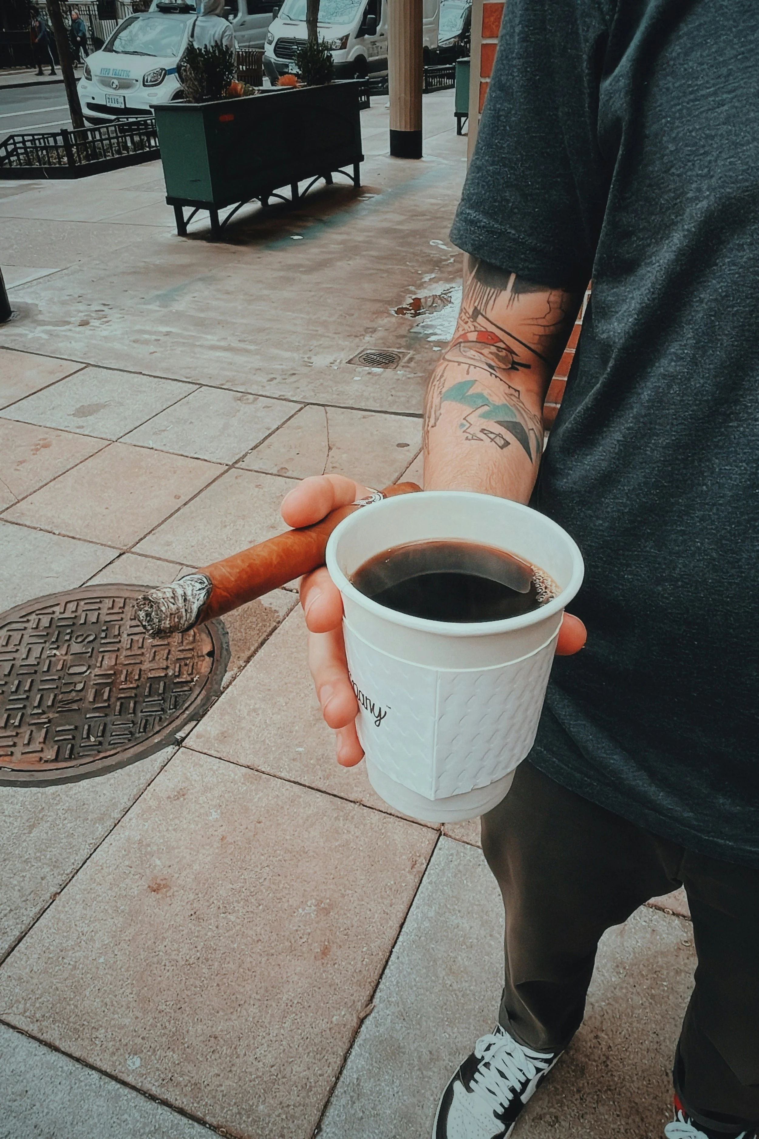 Person holding a white coffee cup in one hand and a cigar in the other, standing on a city sidewalk.