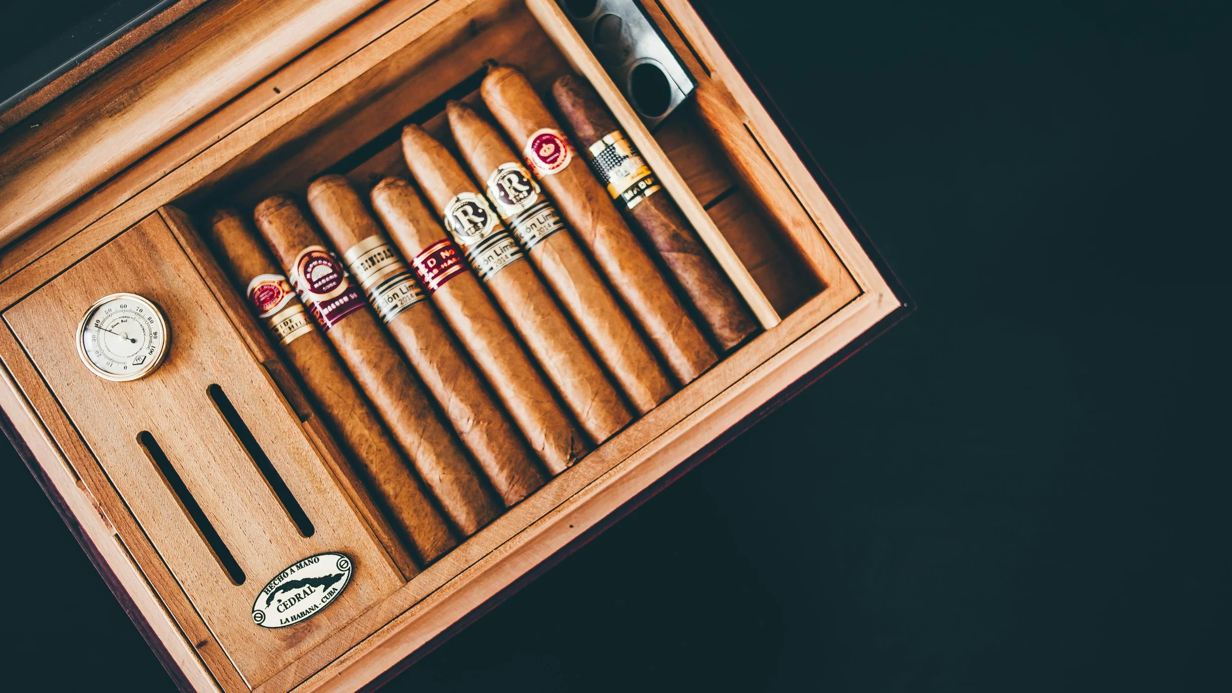 A wooden humidor box containing eight different cigars with labels, a hygrometer on the left, and a CEDAR label on the bottom left.