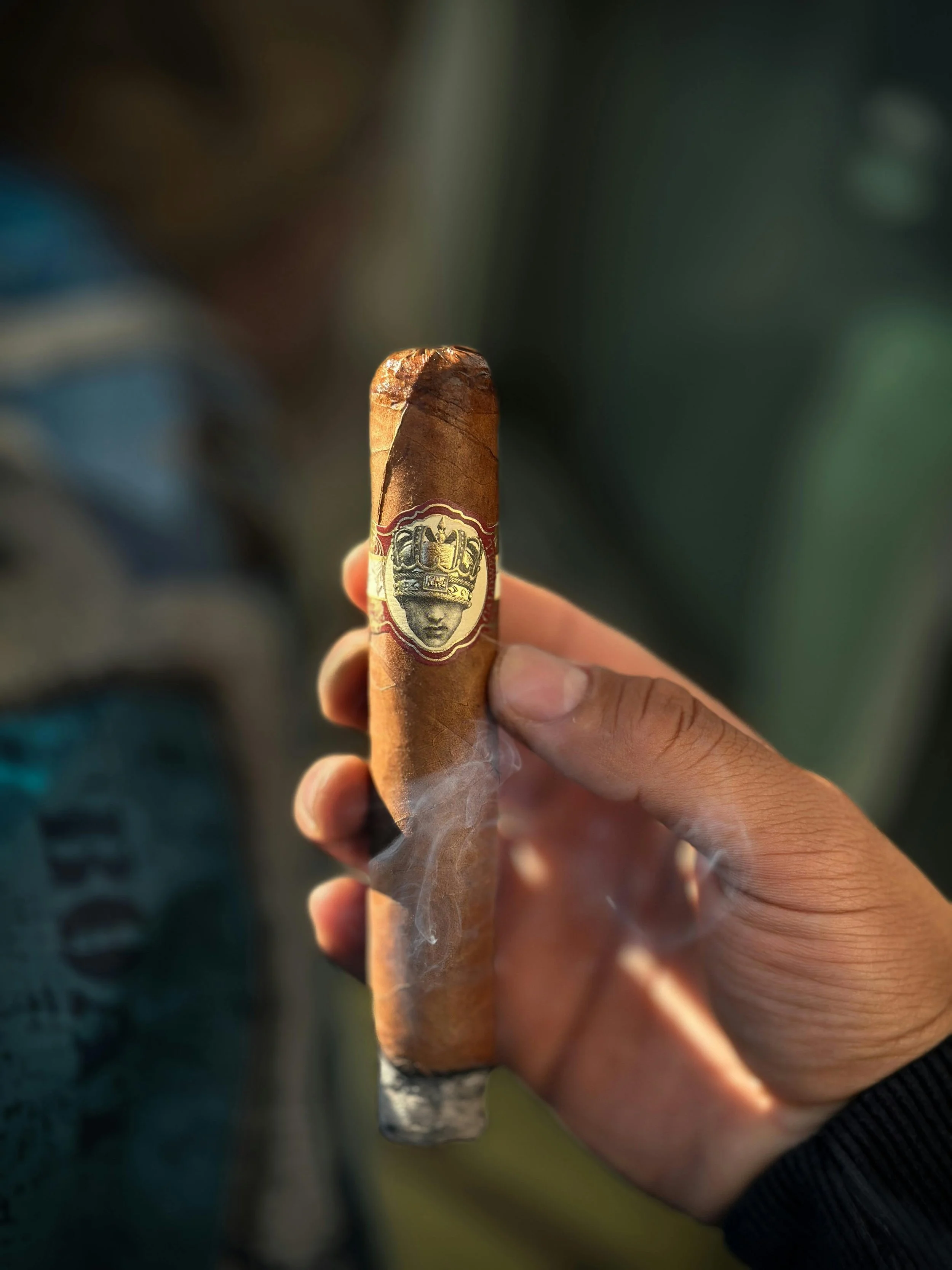 Close-up of a hand holding a lit cigar with a decorative band, with smoke rising from it, against a blurred background.