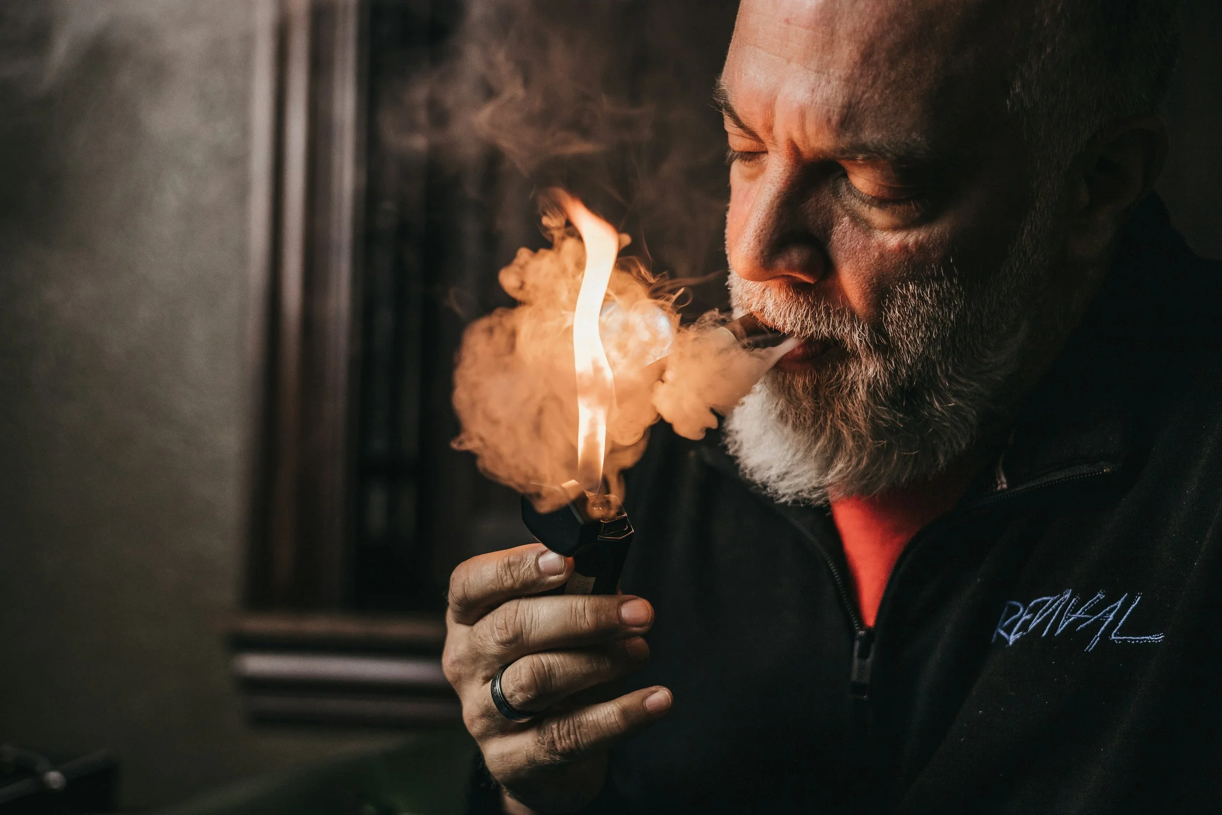 A man with gray hair and beard, wearing a black jacket with embroidered text, is exhaling vapor from an electronic cigarette with a flame visible in the vapor.