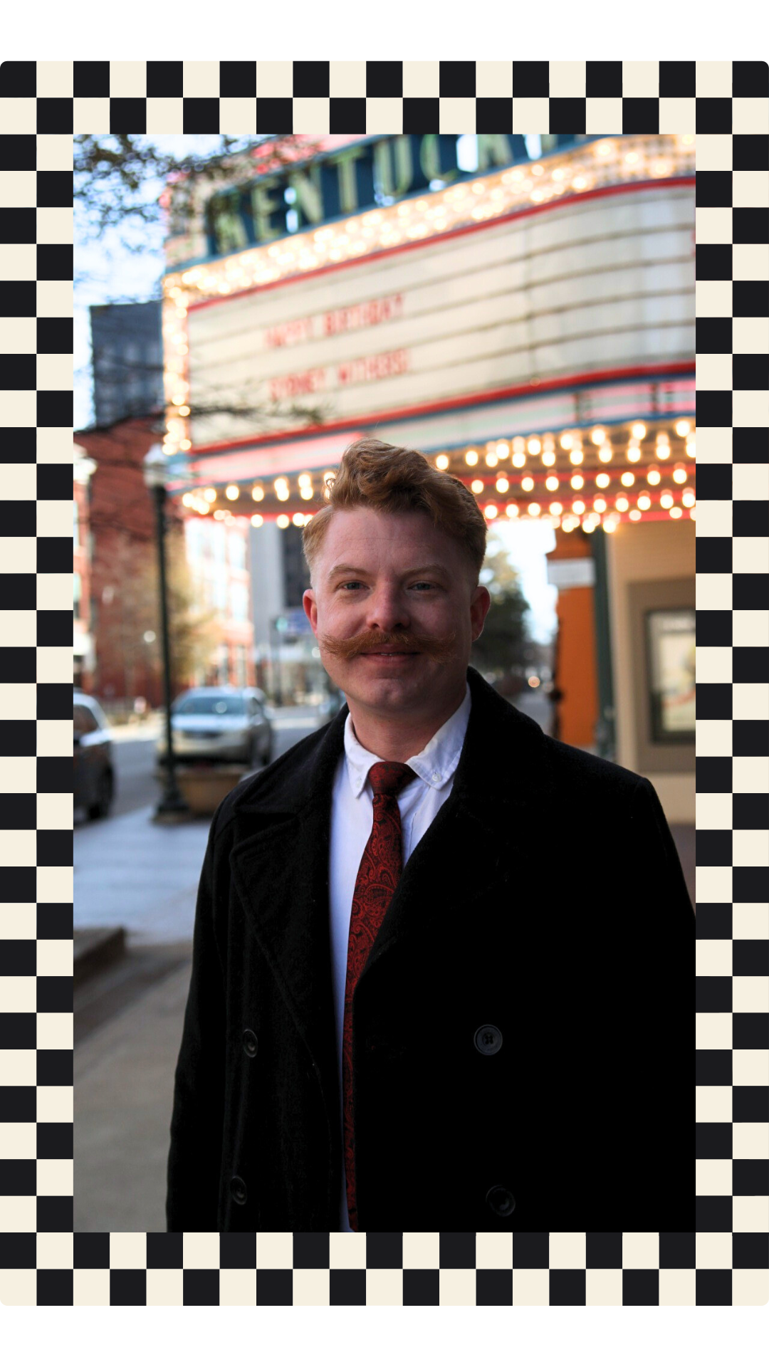 A man with ginger hair, a mustache, and a goatee, dressed in a black coat, white shirt, and red patterned tie, standing on a city sidewalk in front of a vintage theater marquee with lights and a blurred background of cars and buildings.