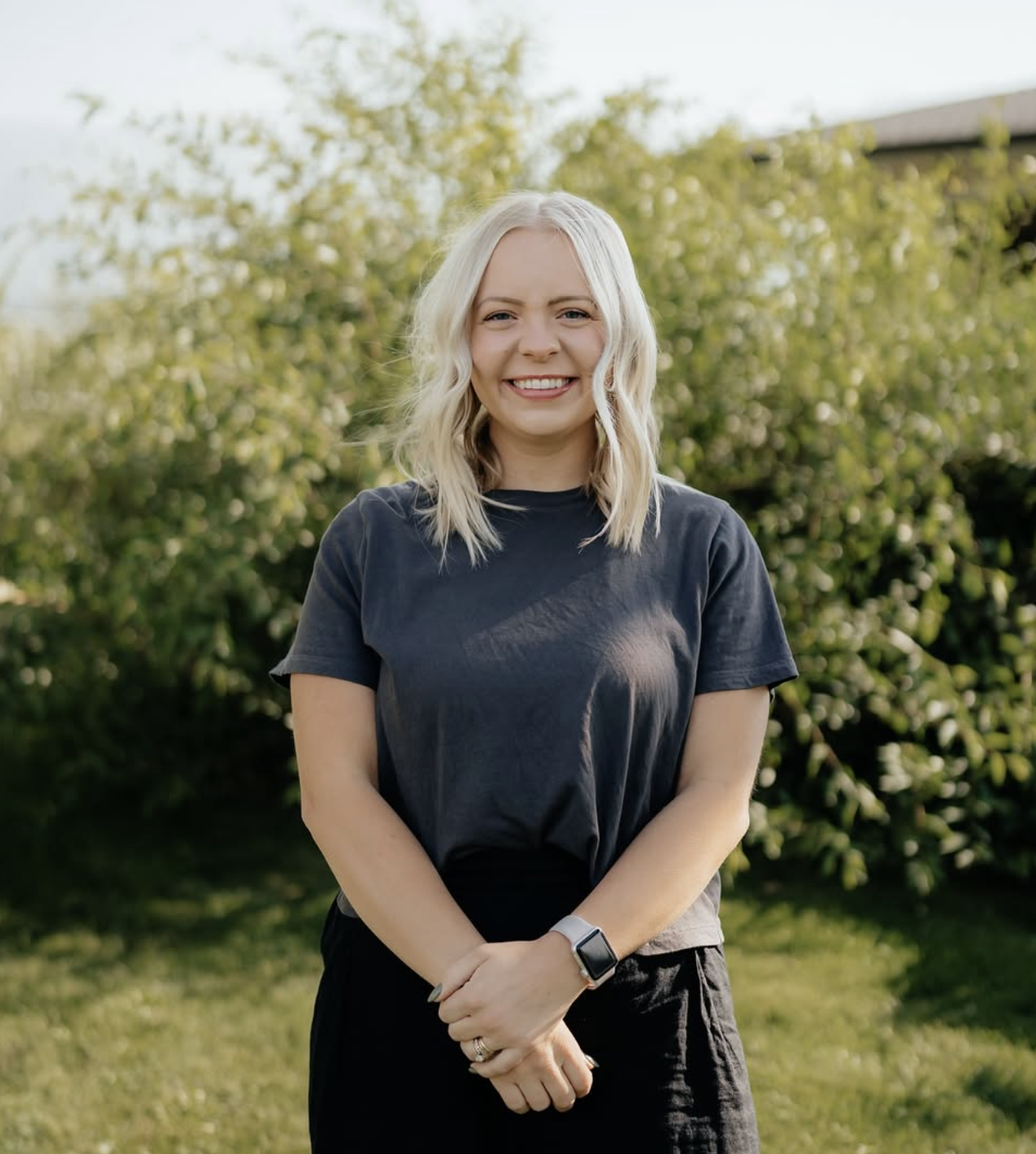 A smiling young woman with wavy blonde hair standing outdoors in front of green bushes and trees, wearing a black t-shirt and a smartwatch on her left wrist.
