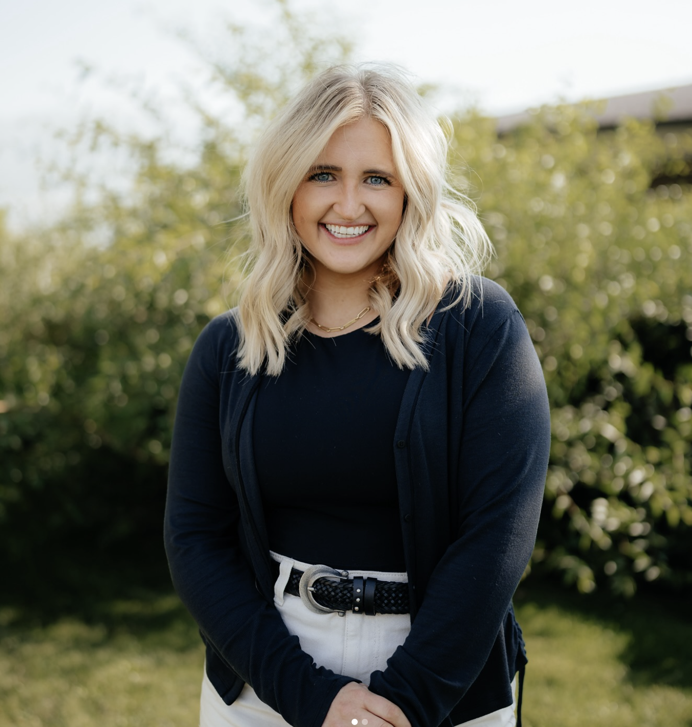 A smiling blonde woman with wavy hair outdoors, dressed in a black top, navy cardigan, and white pants with a black woven belt, standing in a garden with green bushes in the background.