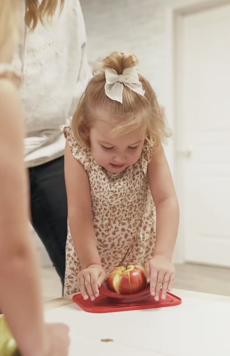 A young girl with a white bow in her hair, wearing a patterned sleeveless dress, looks at a red apple on a plate on a table.