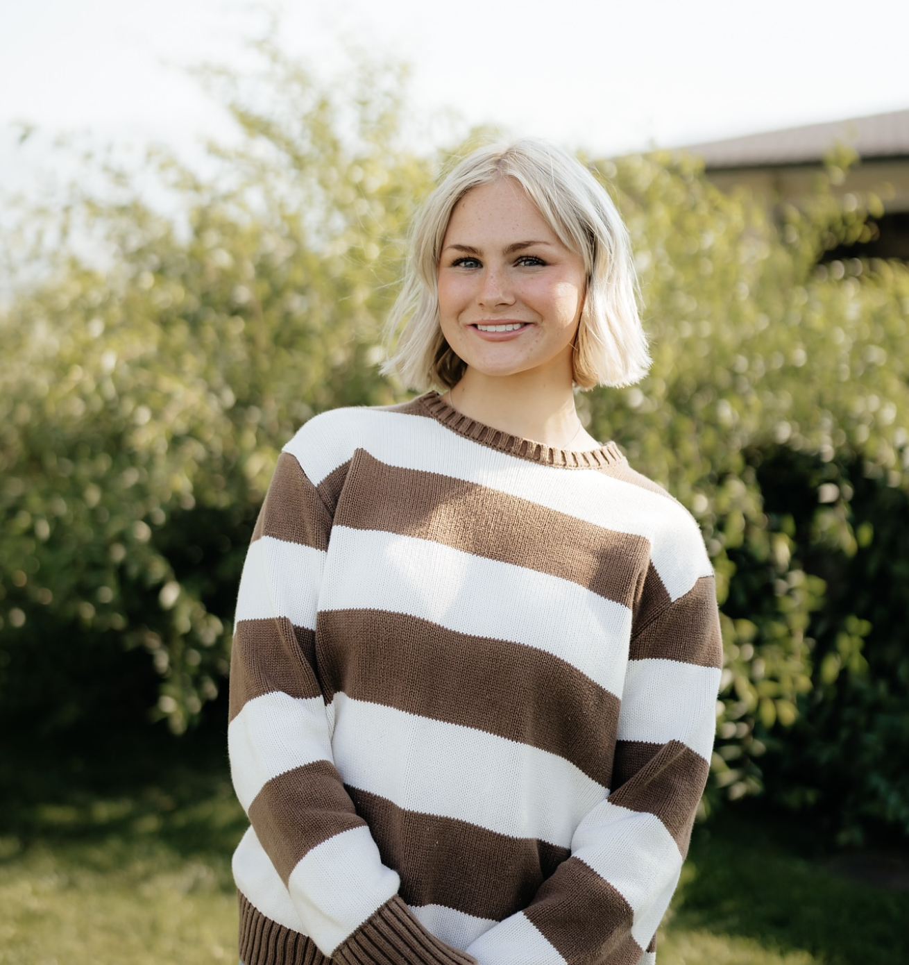 A young woman with blonde hair and a striped brown and white sweater standing outdoors with greenery and trees in the background, smiling