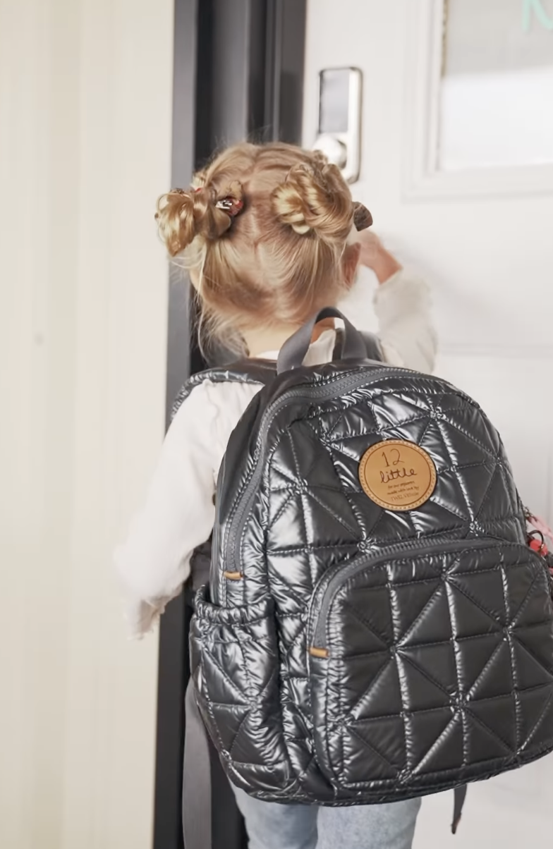 A young girl with blonde hair tied in buns with hair accessories wearing a backpack, standing in front of a door and reaching to open it.